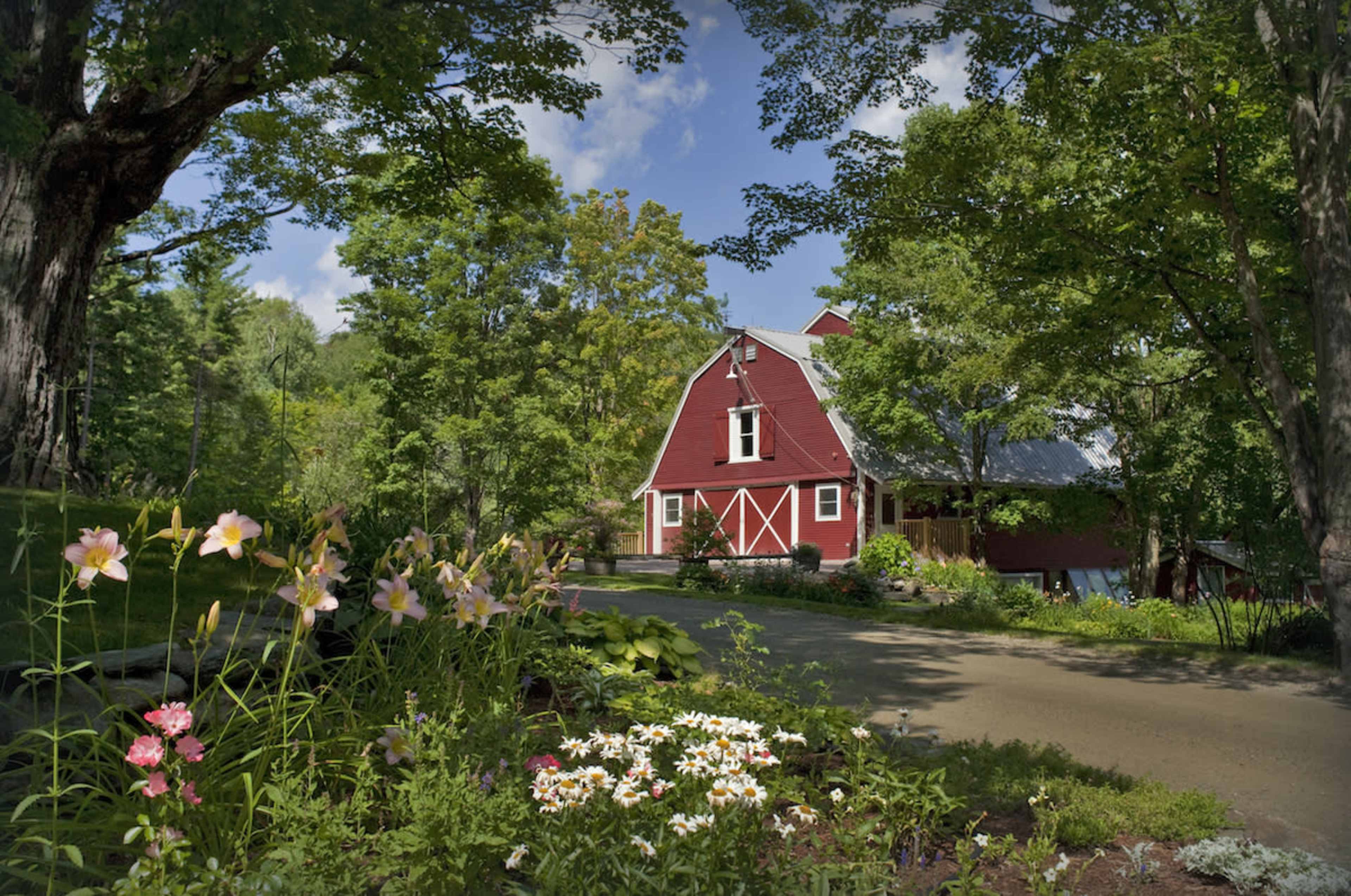 A red barn-style house is surrounded by green trees and blooming flowers along a winding driveway.