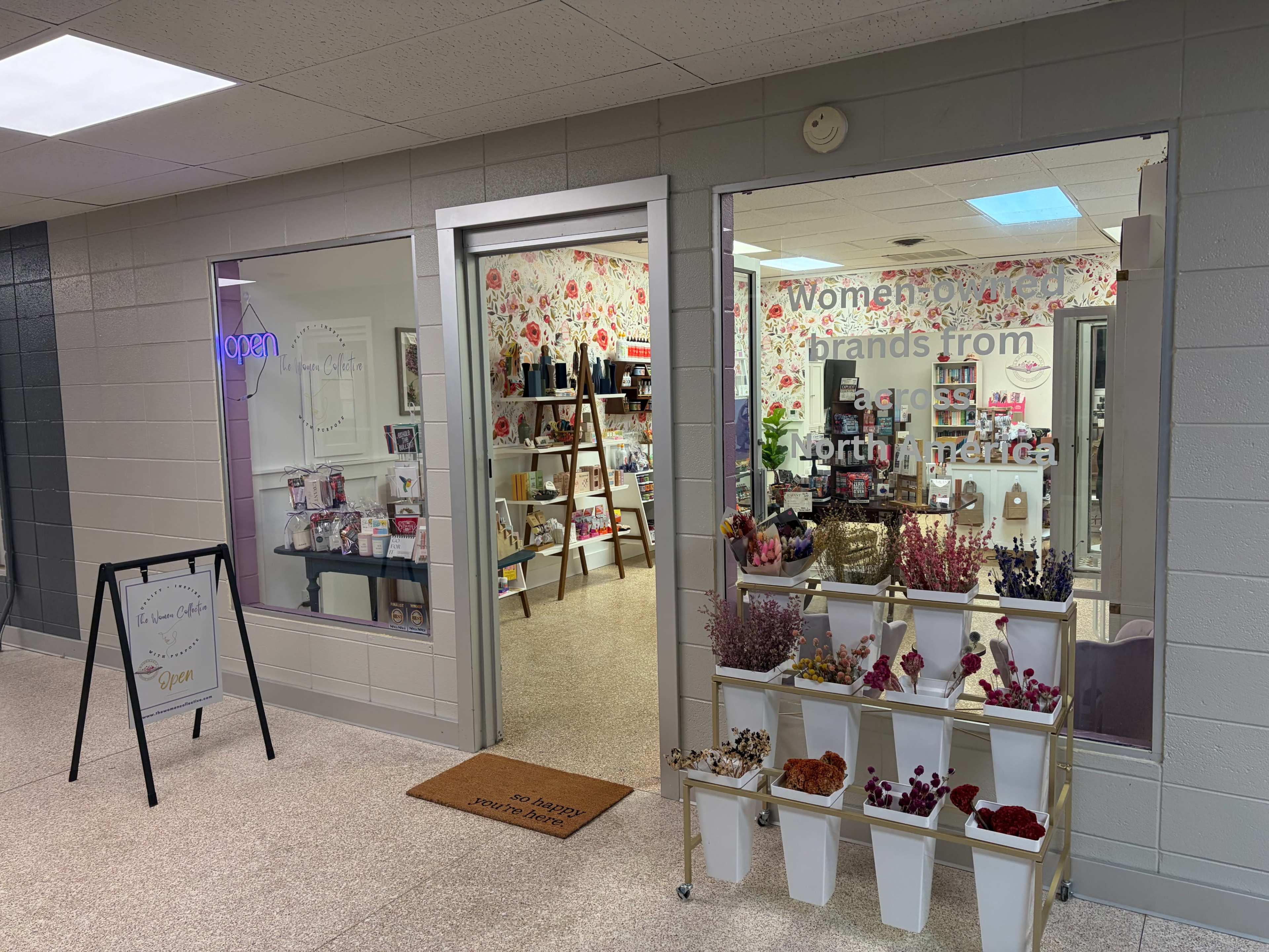 The image shows a retail store display with floral wallpaper and products arranged on shelves, featuring a sign that highlights women-owned brands from North America.