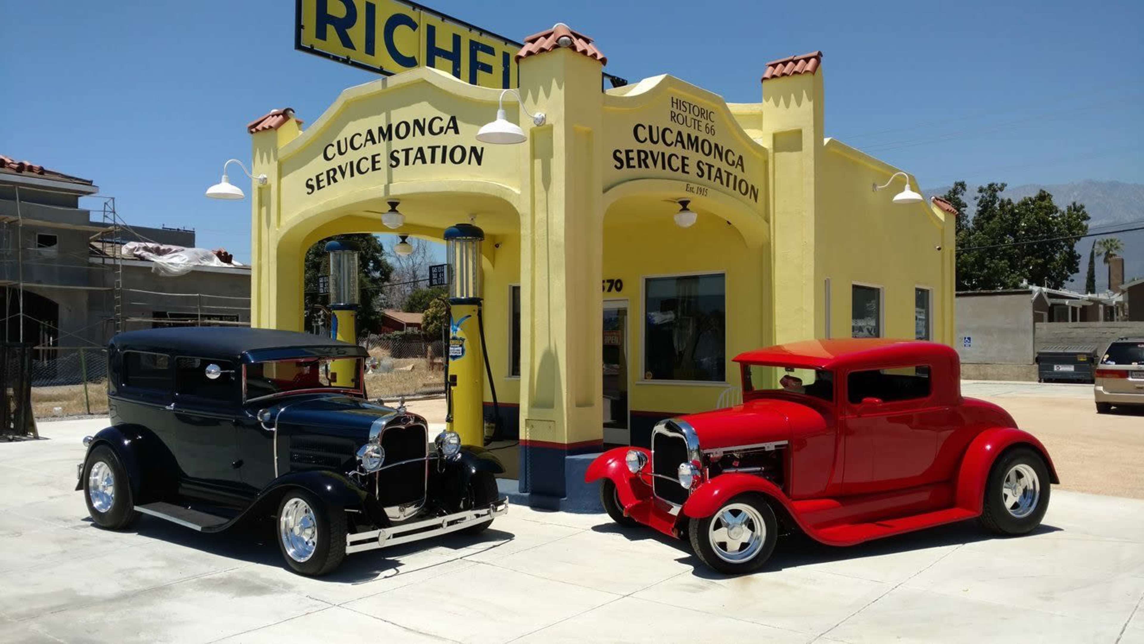 Two vintage cars, one black and one red, park in front of a brightly painted yellow service station with "Cucamonga Service Station" signage.