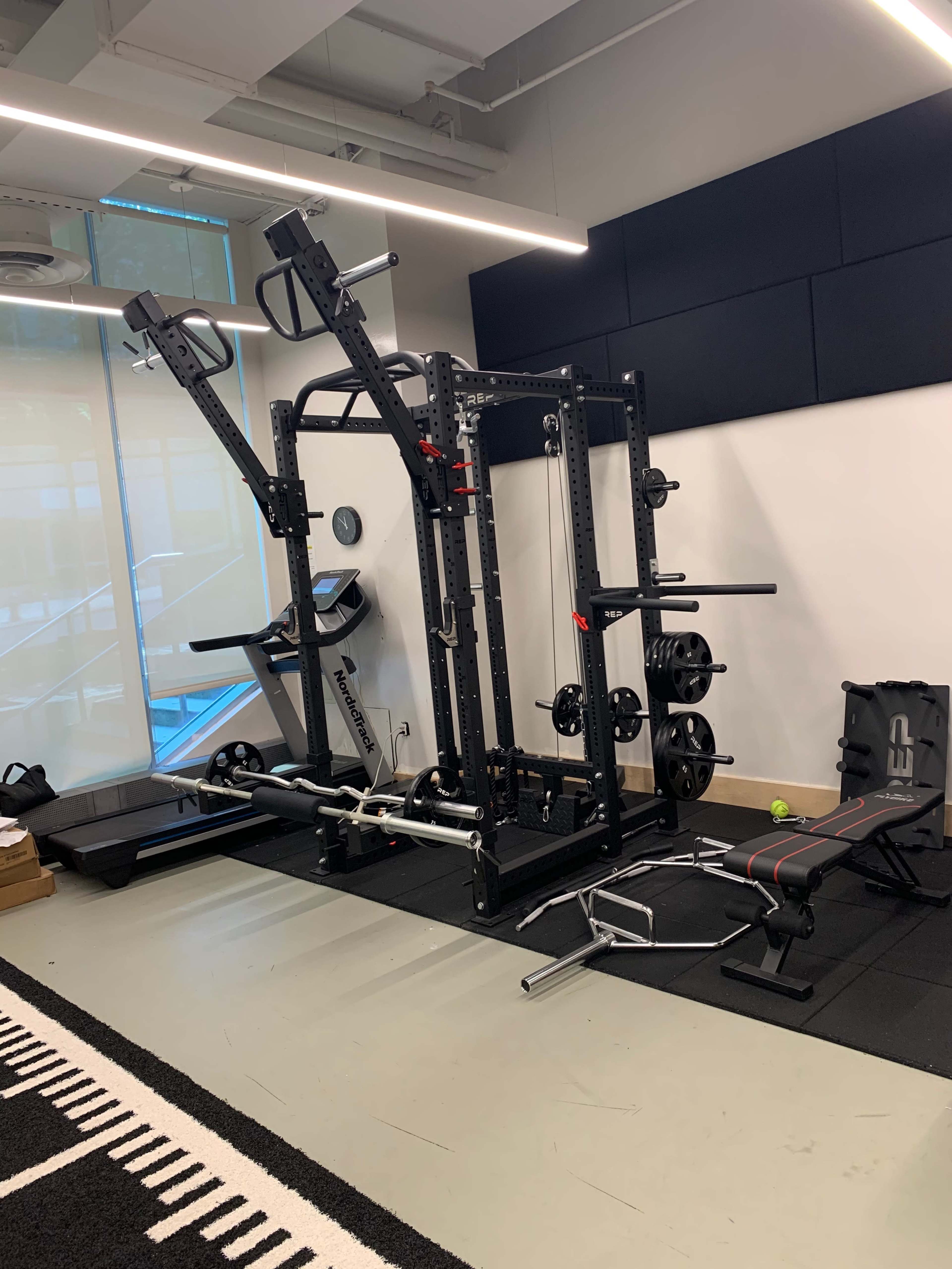 The image shows a home gym equipped with a power rack, weights, a treadmill, and a bench on a rubber mat.