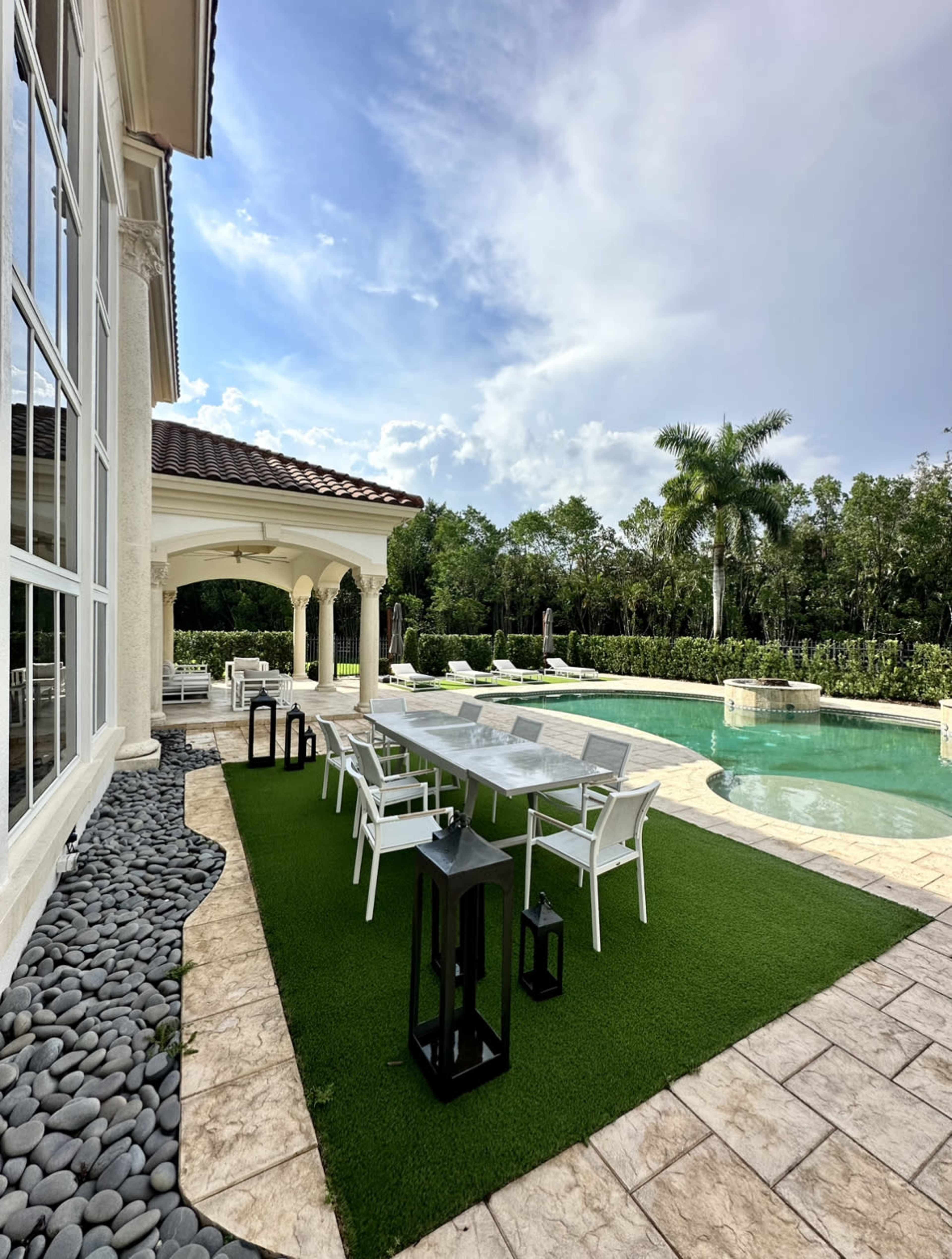 A patio area with a long dining table and chairs beside a swimming pool, surrounded by greenery and decorative lanterns.