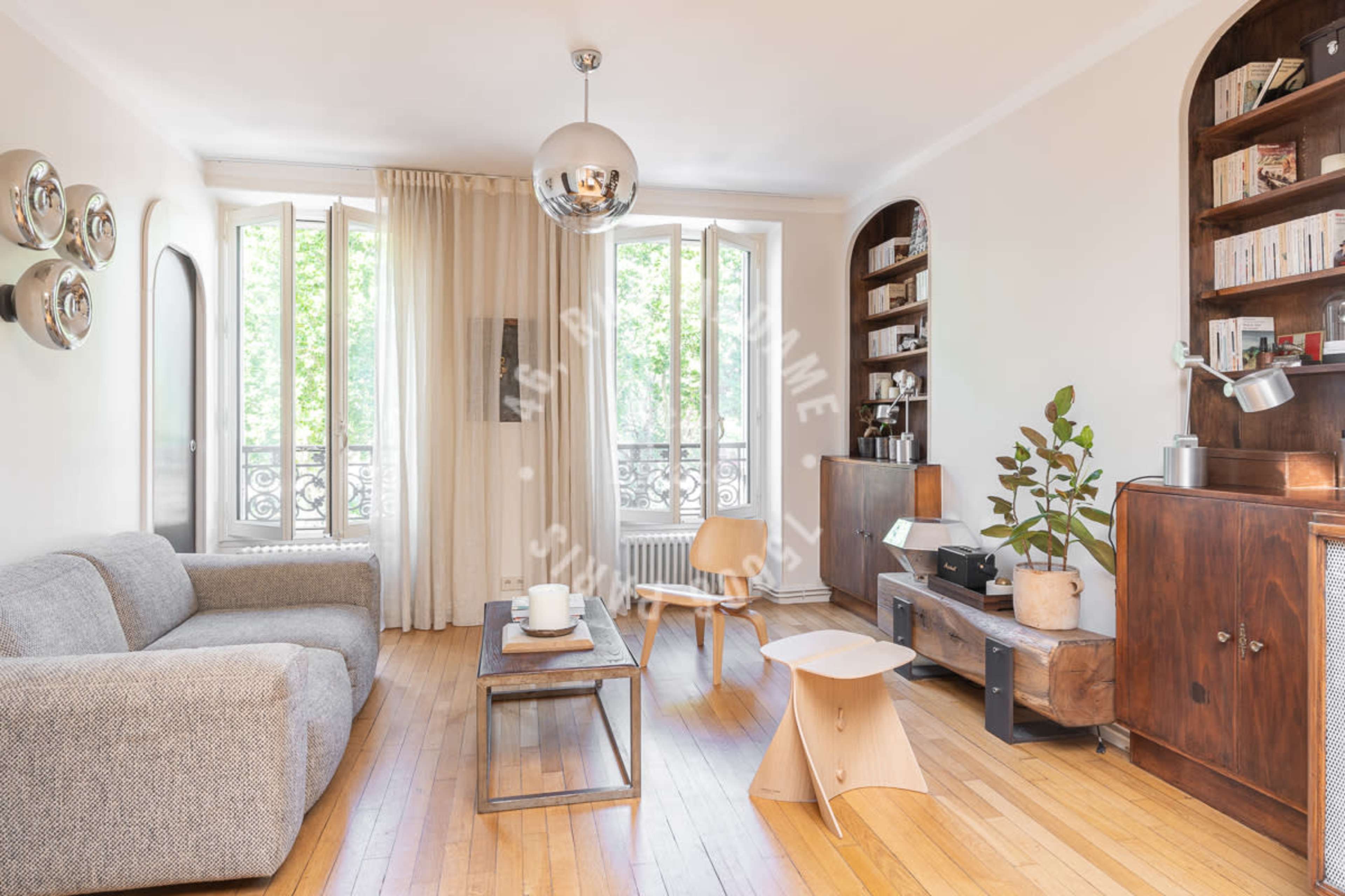 The living room features a gray sofa, a wooden coffee table, and shelves with various items, illuminated by natural light from large windows.