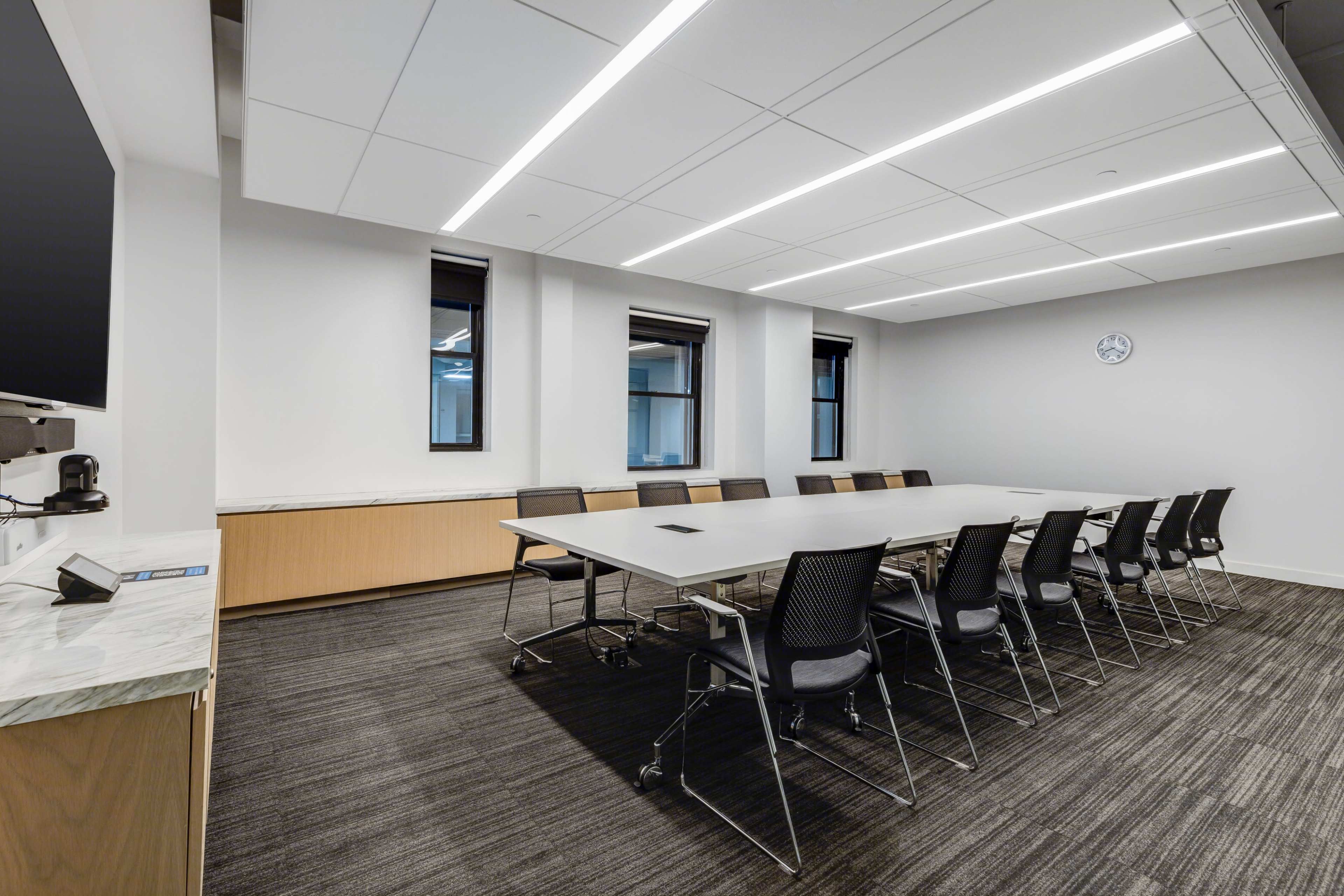 A conference room features a long table surrounded by chairs, with large windows and overhead lighting.