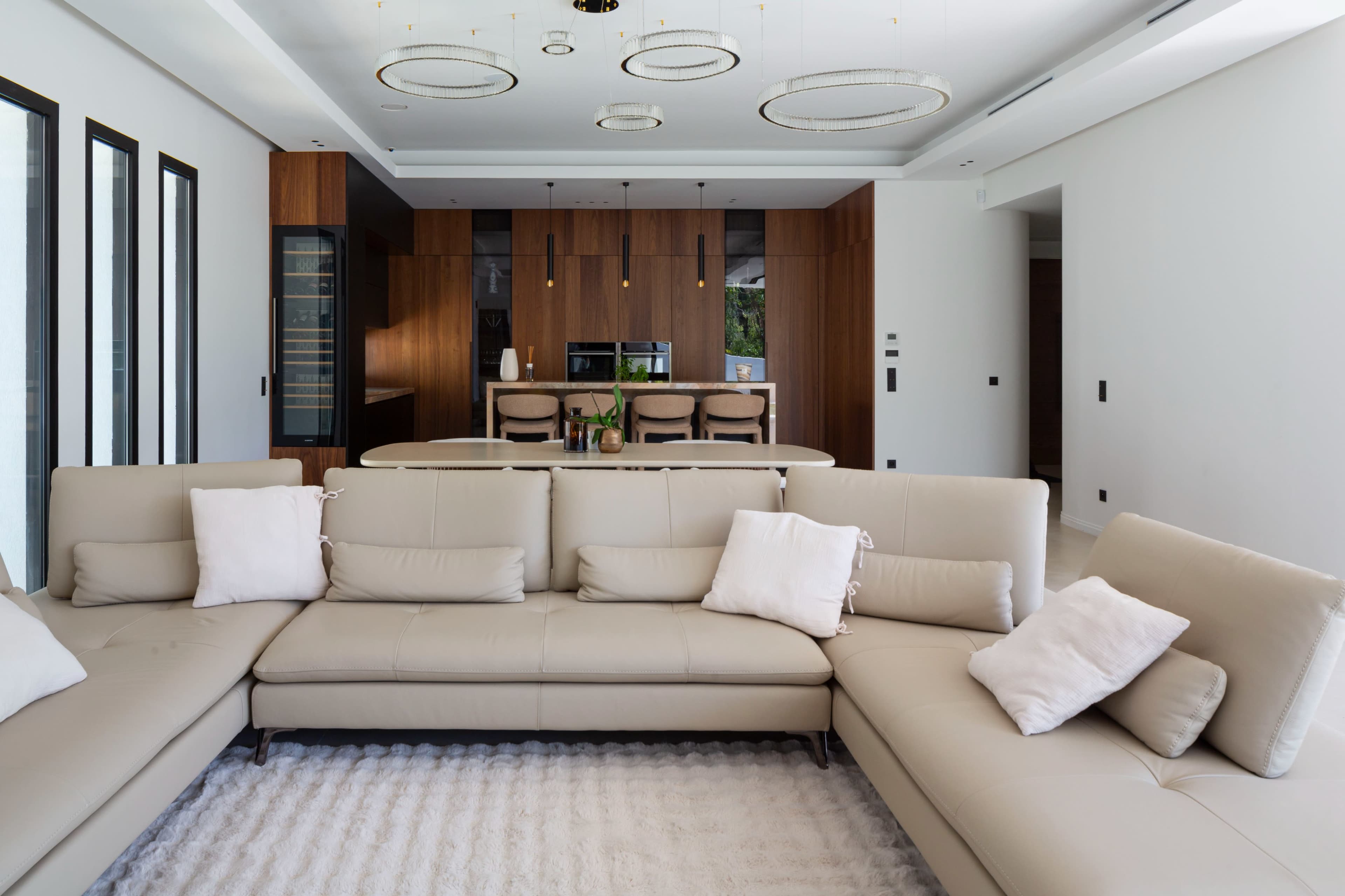 A modern living room features a large beige sectional sofa with several pillows, set against a backdrop of a sleek kitchen with wooden cabinetry and a central island.