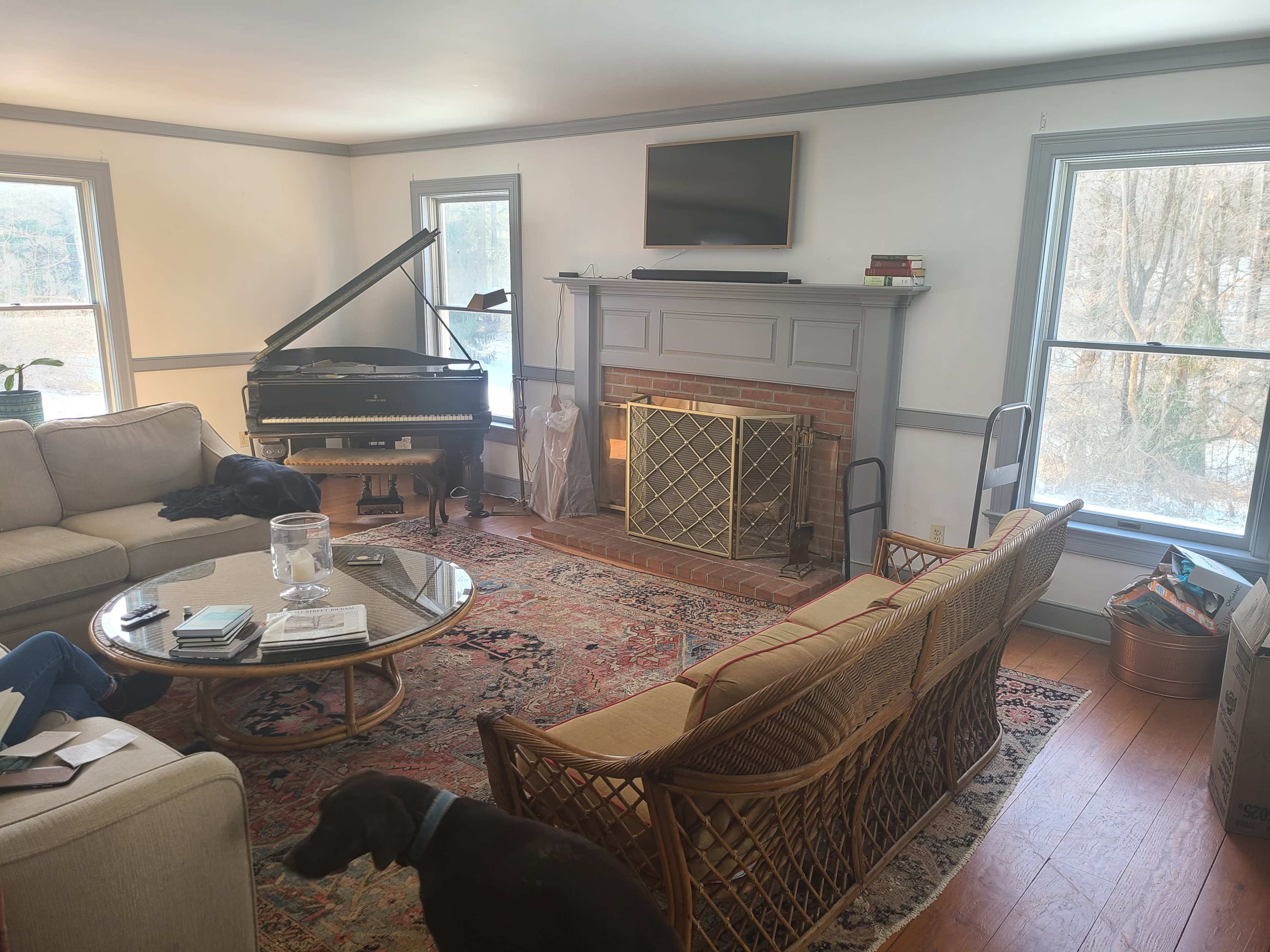The image shows a cozy living room with a grand piano, a seating area featuring a light-colored sofa and a round coffee table, a fireplace, and large windows letting in natural light.