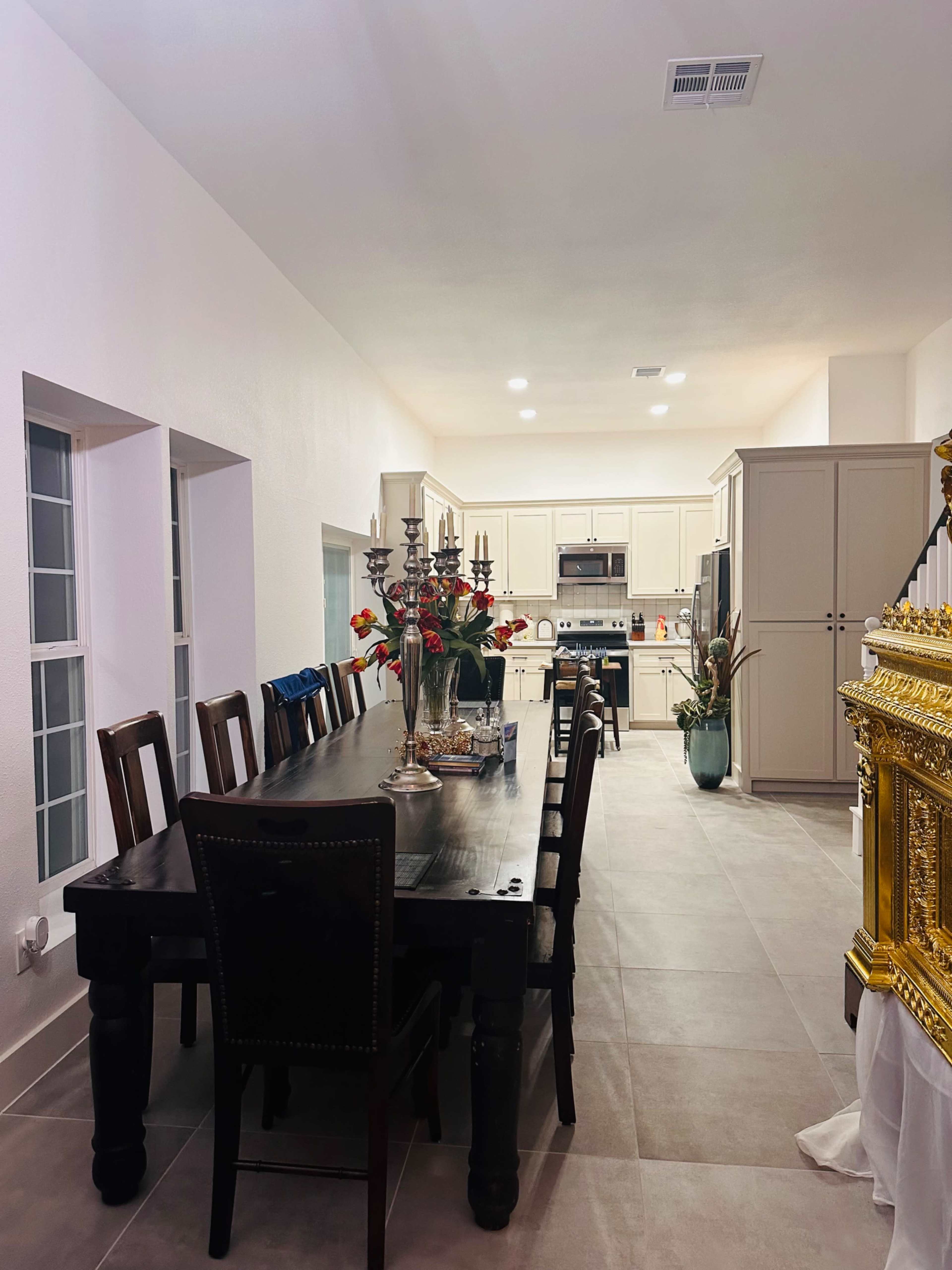 A spacious kitchen and dining area features a long wooden table surrounded by chairs, with white cabinets and large windows providing ample light.