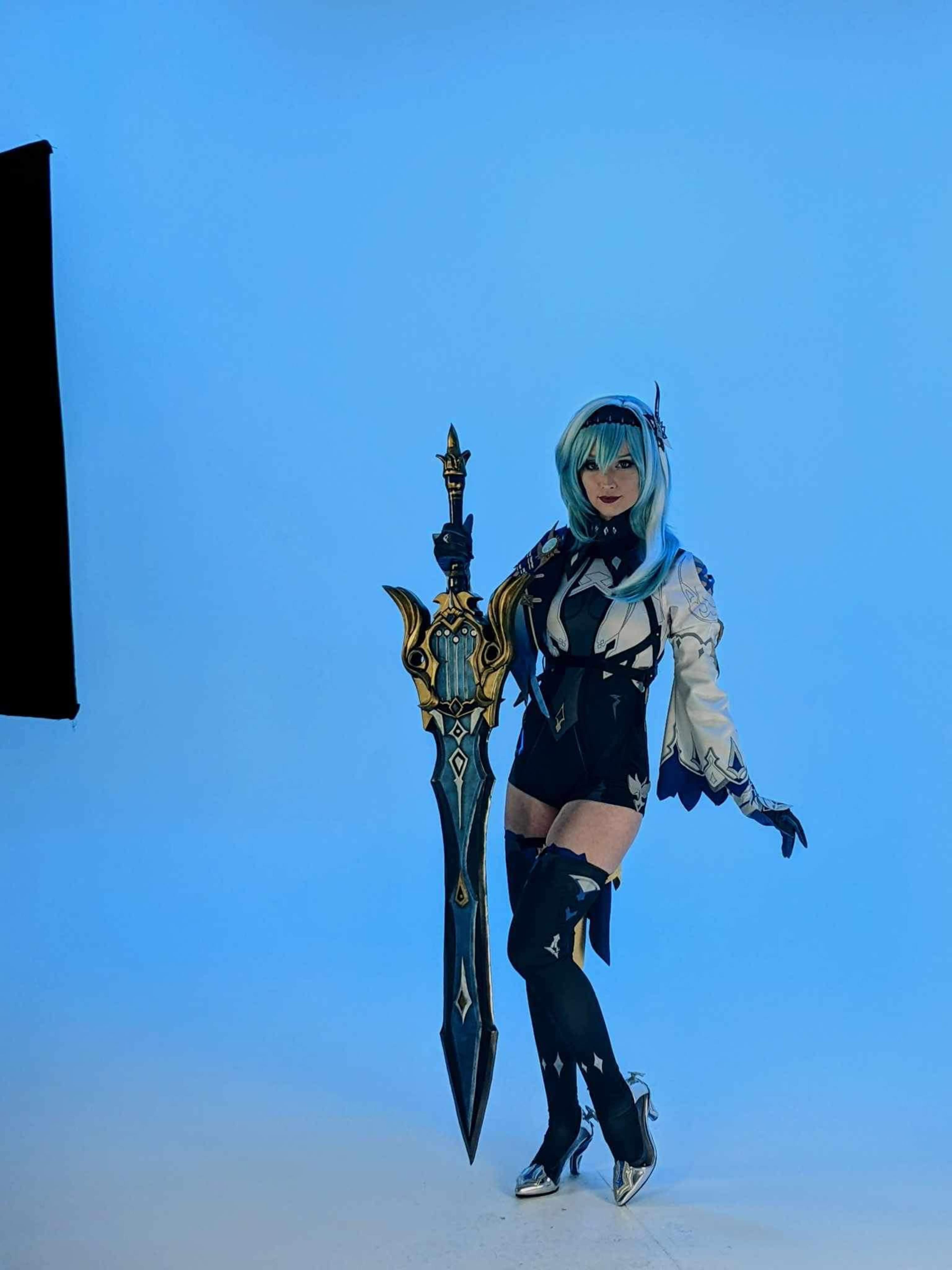 A person poses with a large, ornate sword against a blue background, wearing a detailed costume with a mix of white and black.