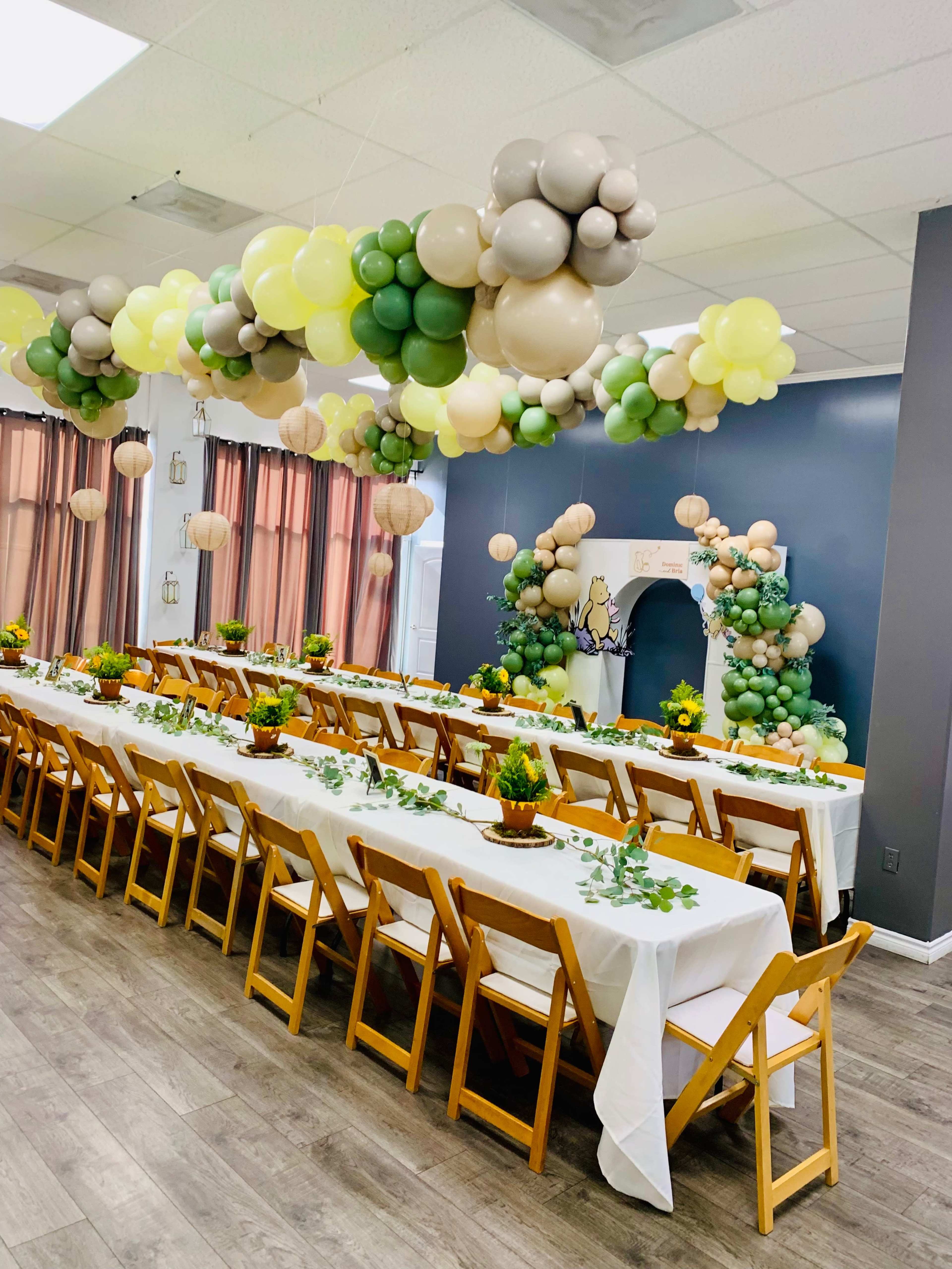 A spacious event room features long tables set with white tablecloths, surrounded by wooden chairs, and adorned with balloon decorations in various shades hanging from the ceiling.