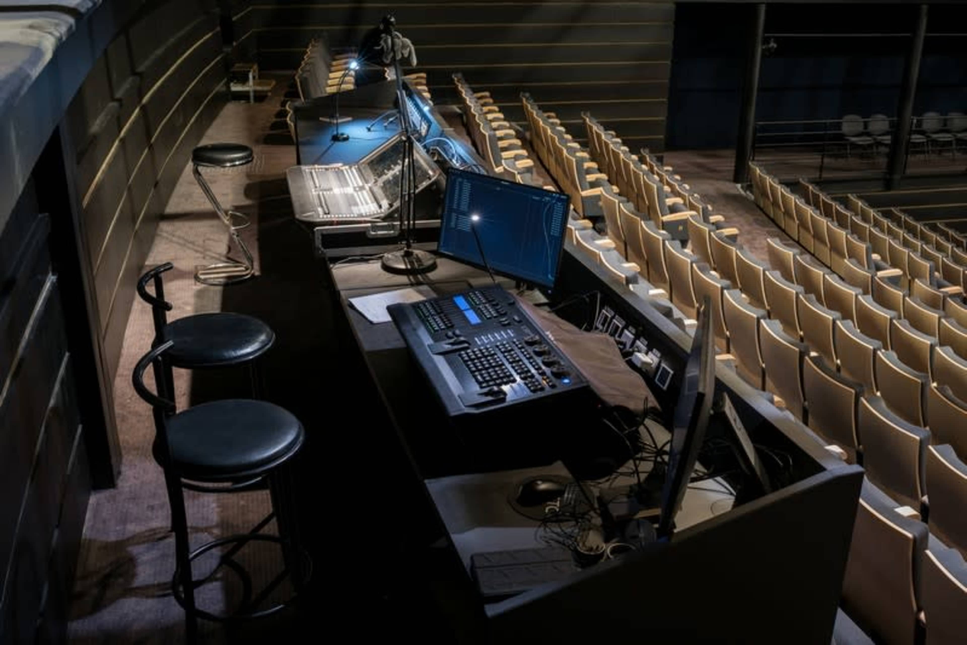 The image shows a dimly lit auditorium with empty rows of seats and a sound or lighting control station featuring multiple screens and equipment.