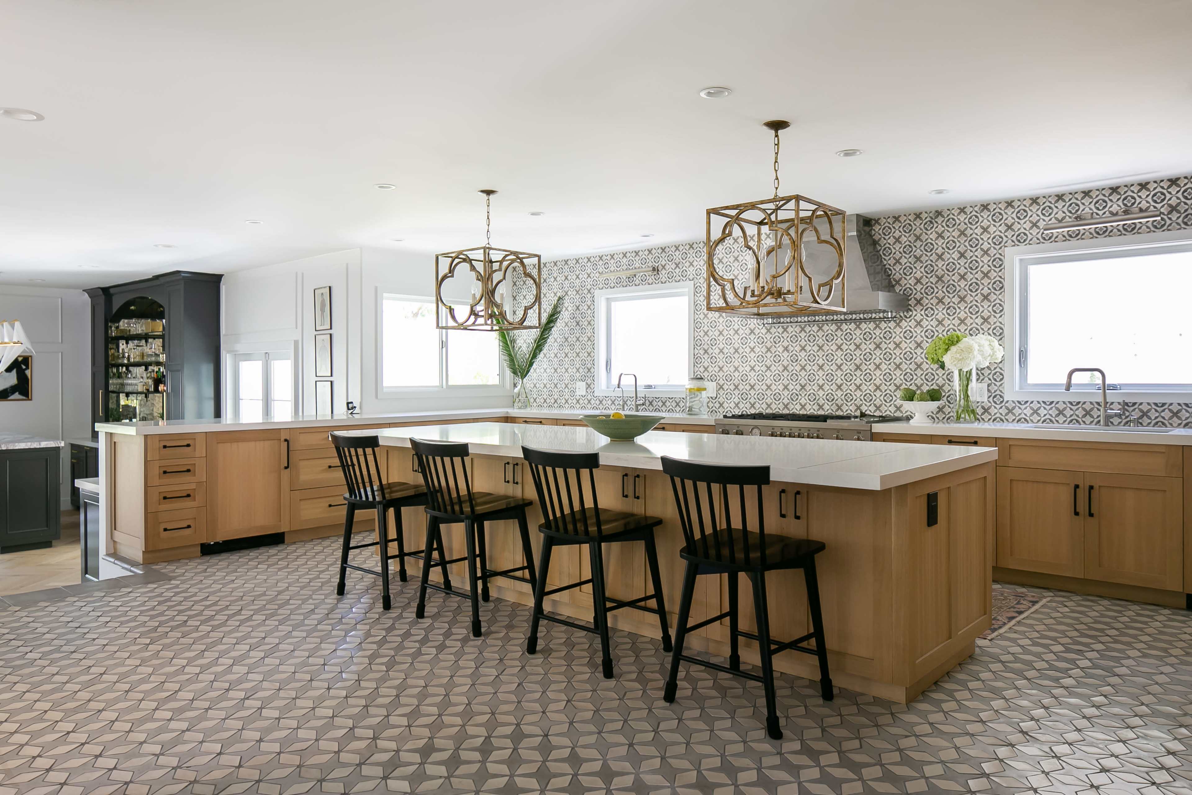 A spacious kitchen features an island with black chairs, light wooden cabinetry, and patterned tile flooring.