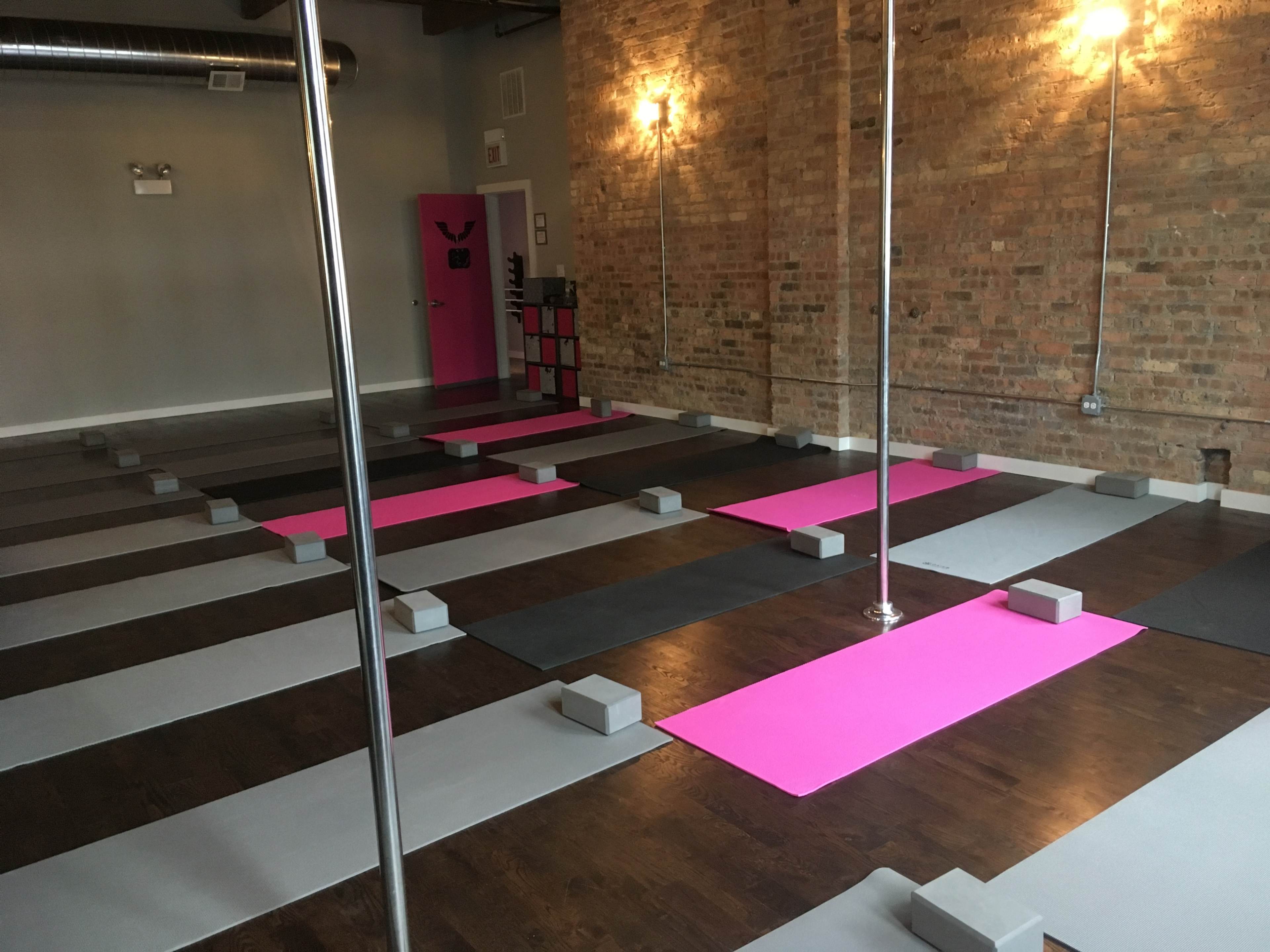 A fitness studio with multiple exercise mats arranged in rows, two pole dance poles, and yoga blocks placed on a wooden floor.