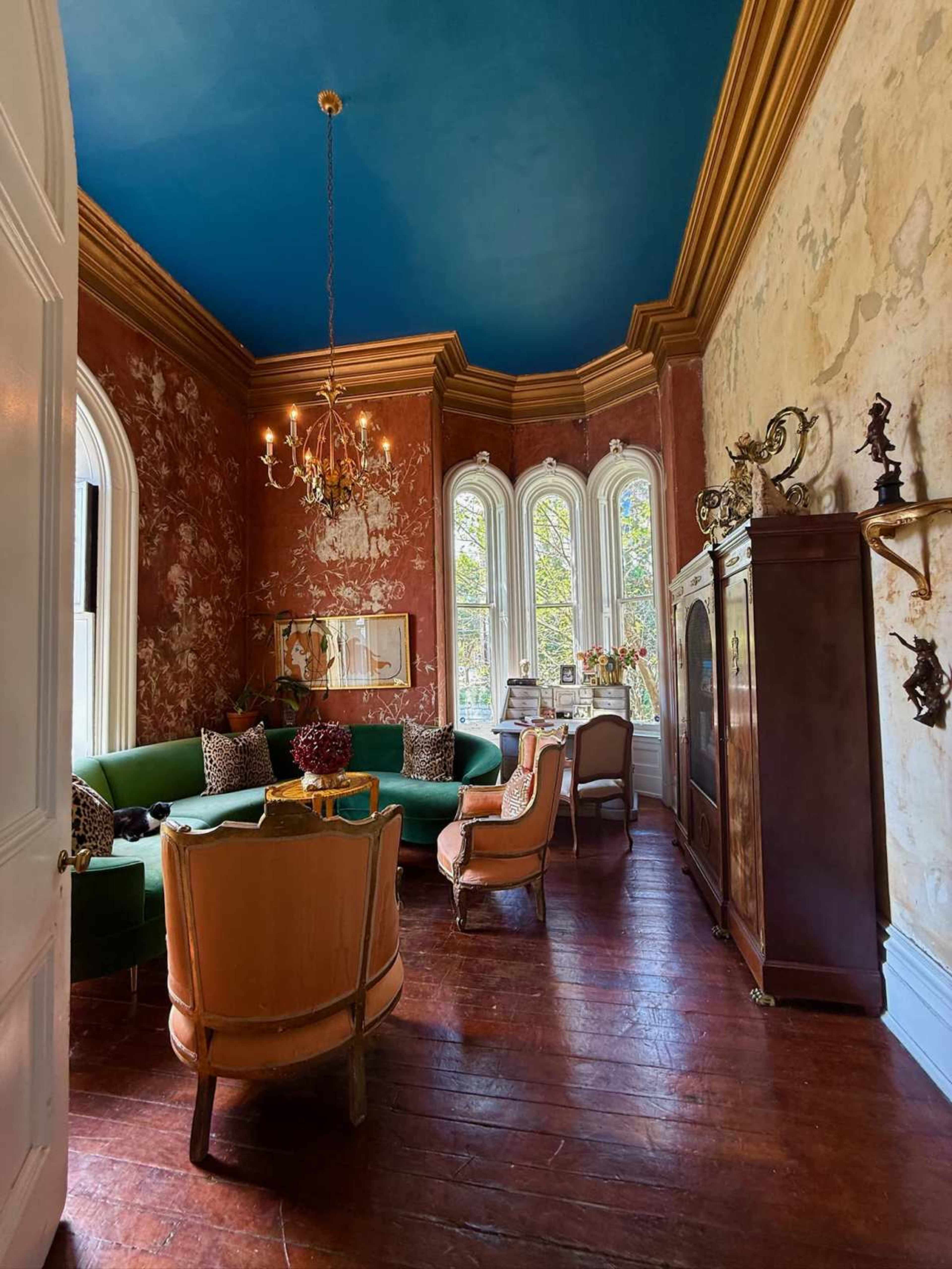 The image shows a cozy sitting room with a green velvet sofa, vintage armchairs, tall windows, and ornate wall decorations featuring a blue painted ceiling.