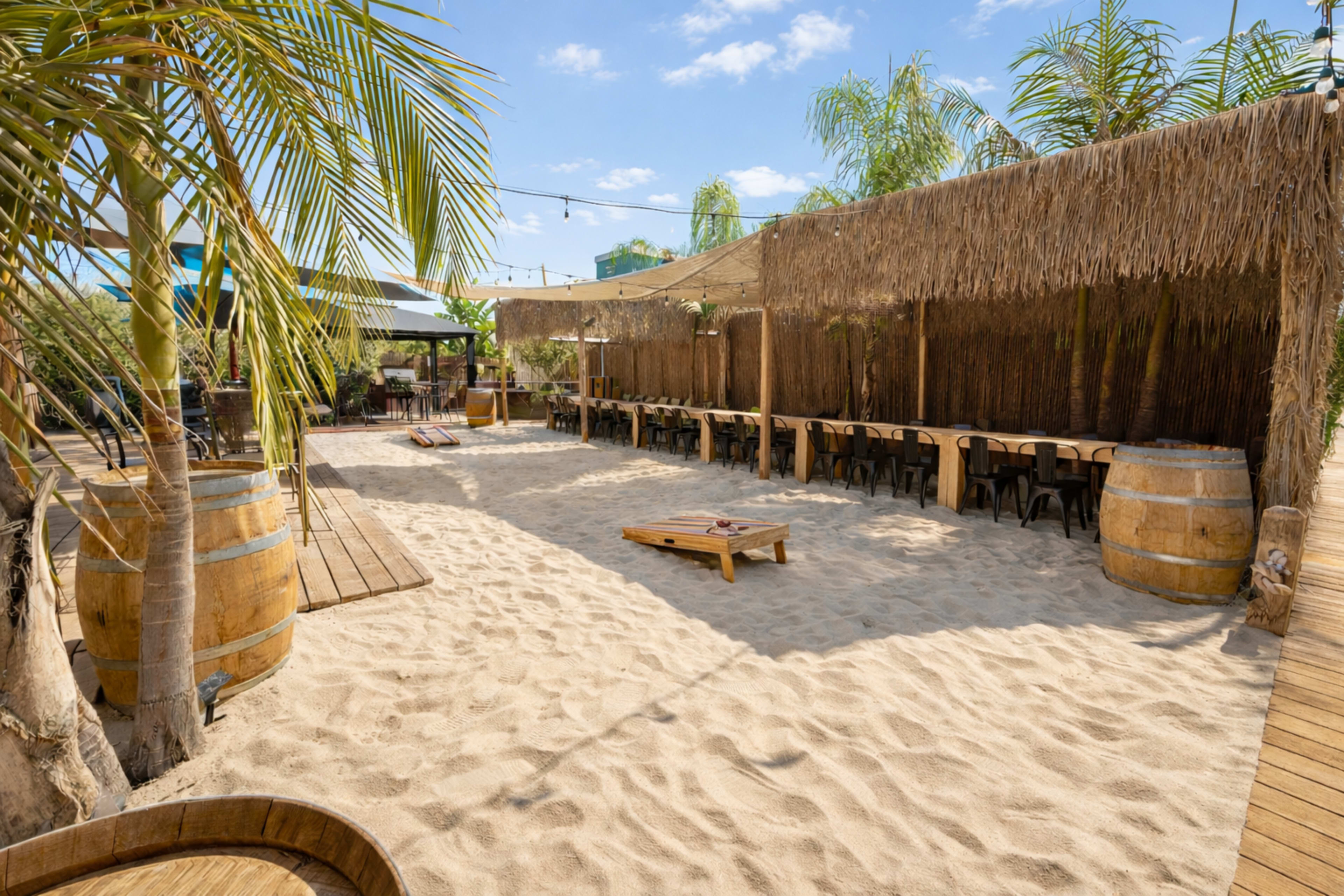 The image shows a sandy outdoor seating area with wooden tables, palm trees, and thatched roof structures.