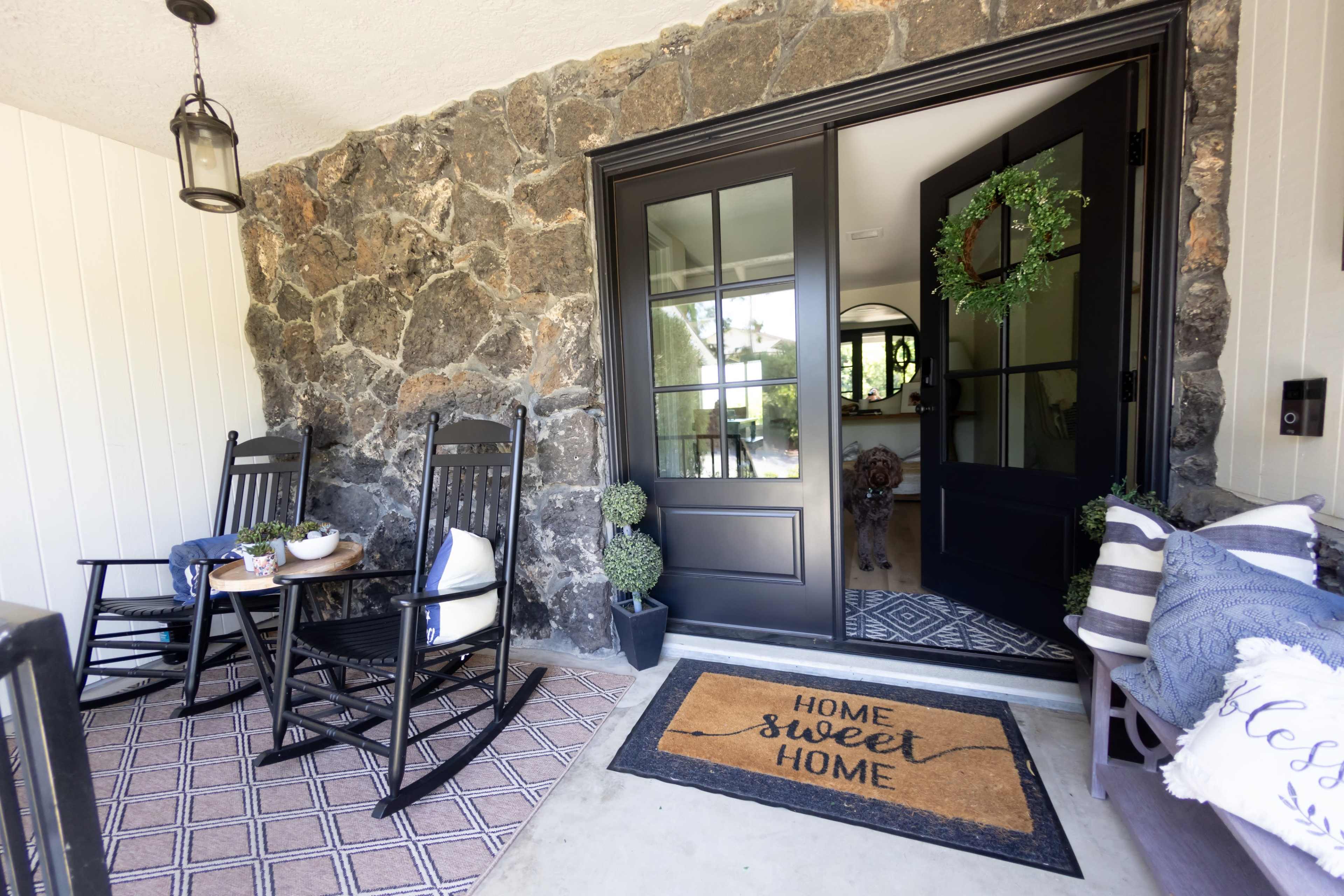 The image shows a charming entryway with a stone wall, a welcoming "Home Sweet Home" doormat, two rocking chairs, and a view into a well-lit interior.