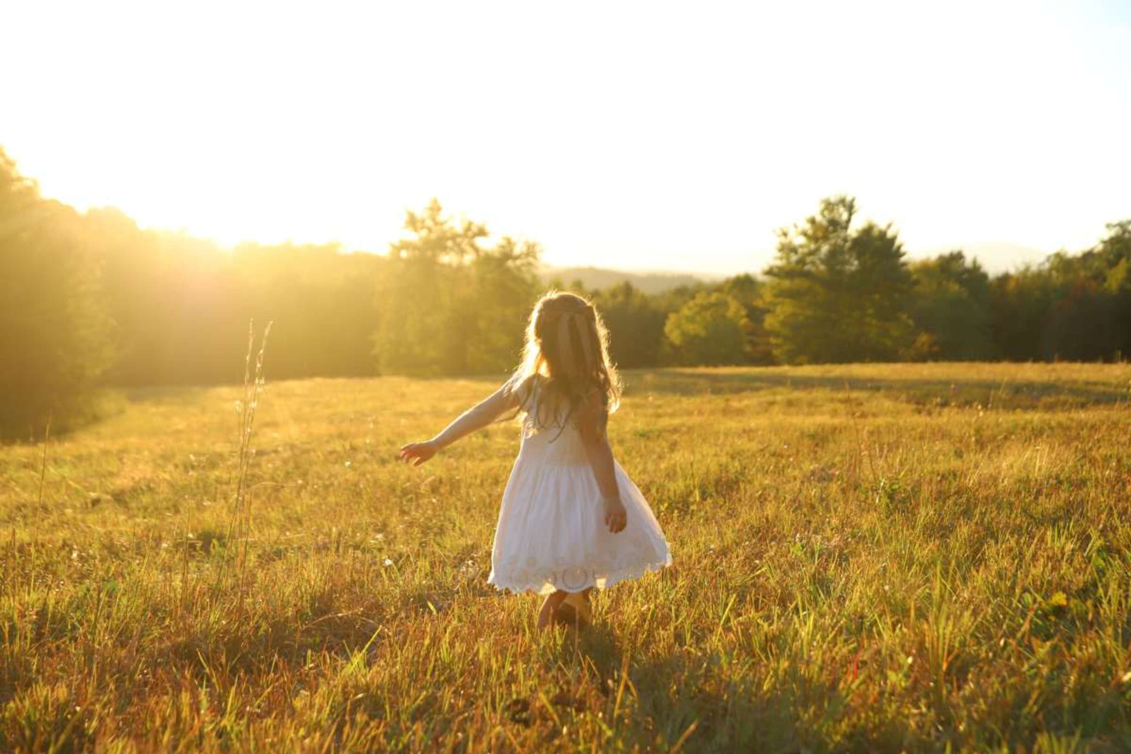 A girl in a white dress walks through a grassy field as the sun sets in the background.