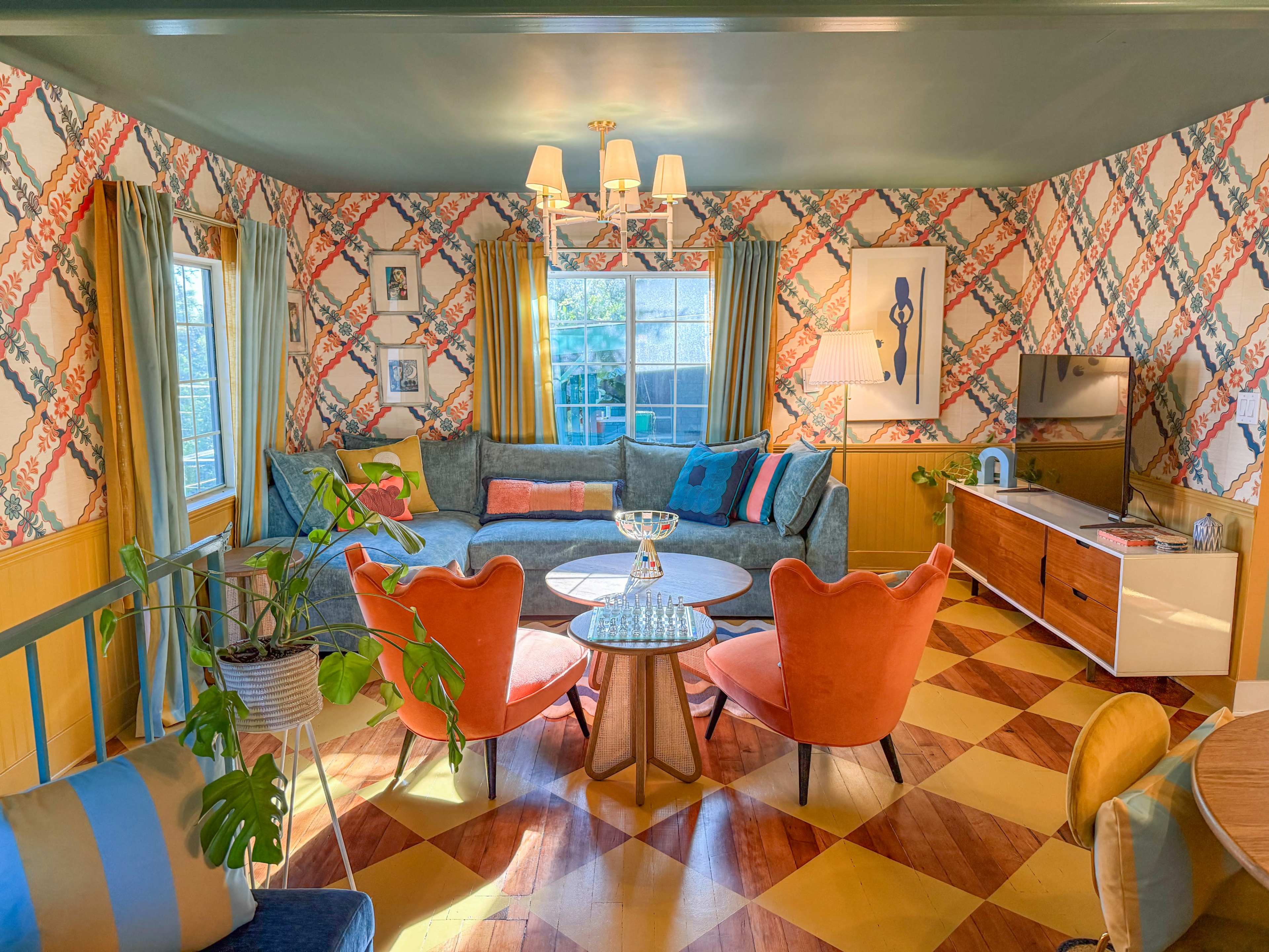 The image shows a colorful living room with patterned wallpaper, a large sectional sofa, round coffee table, and mid-century modern furniture elements.