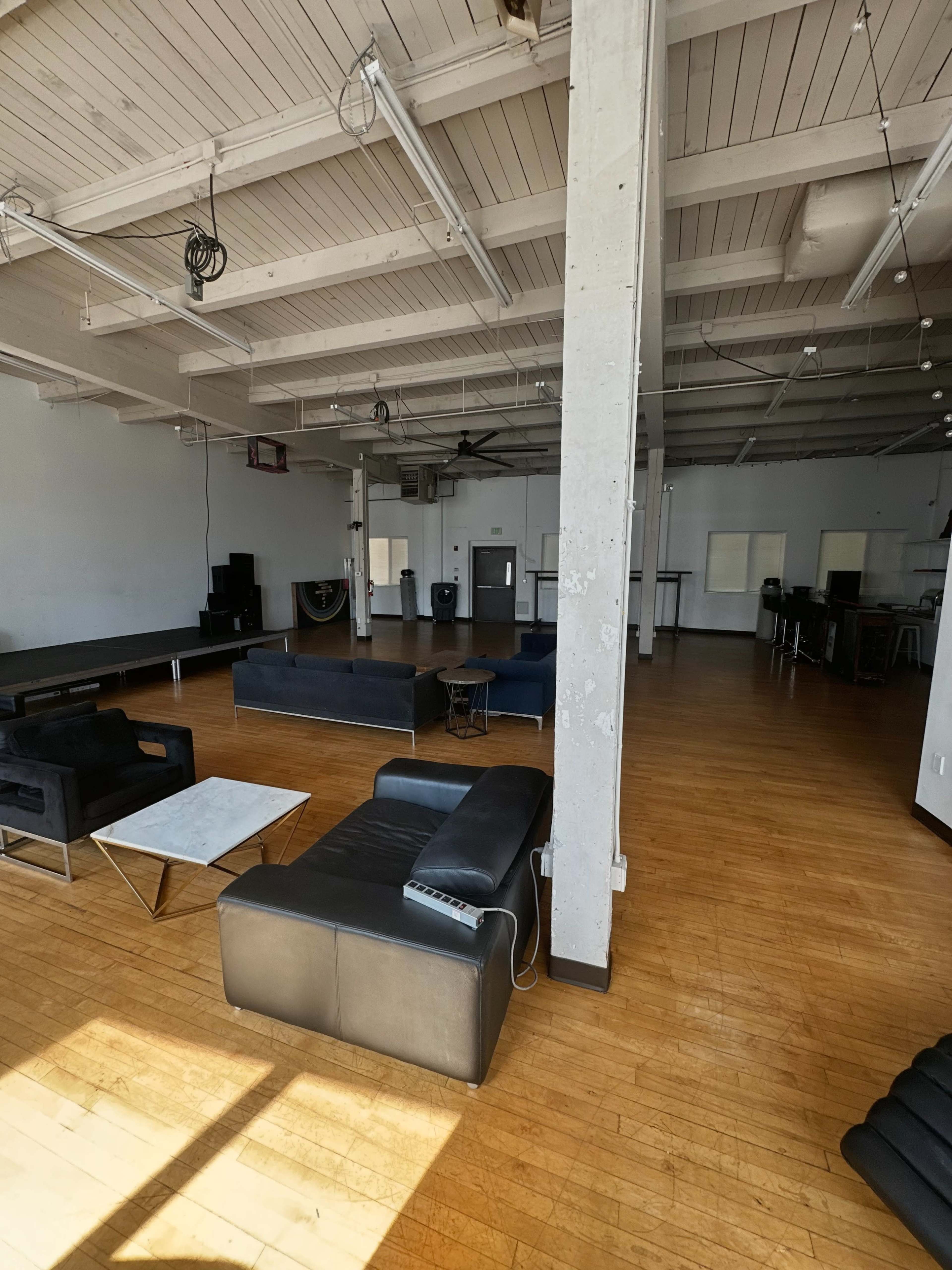 The image shows a spacious, empty room with wooden floors, high ceilings, and black furniture arranged, including sofas and a coffee table.
