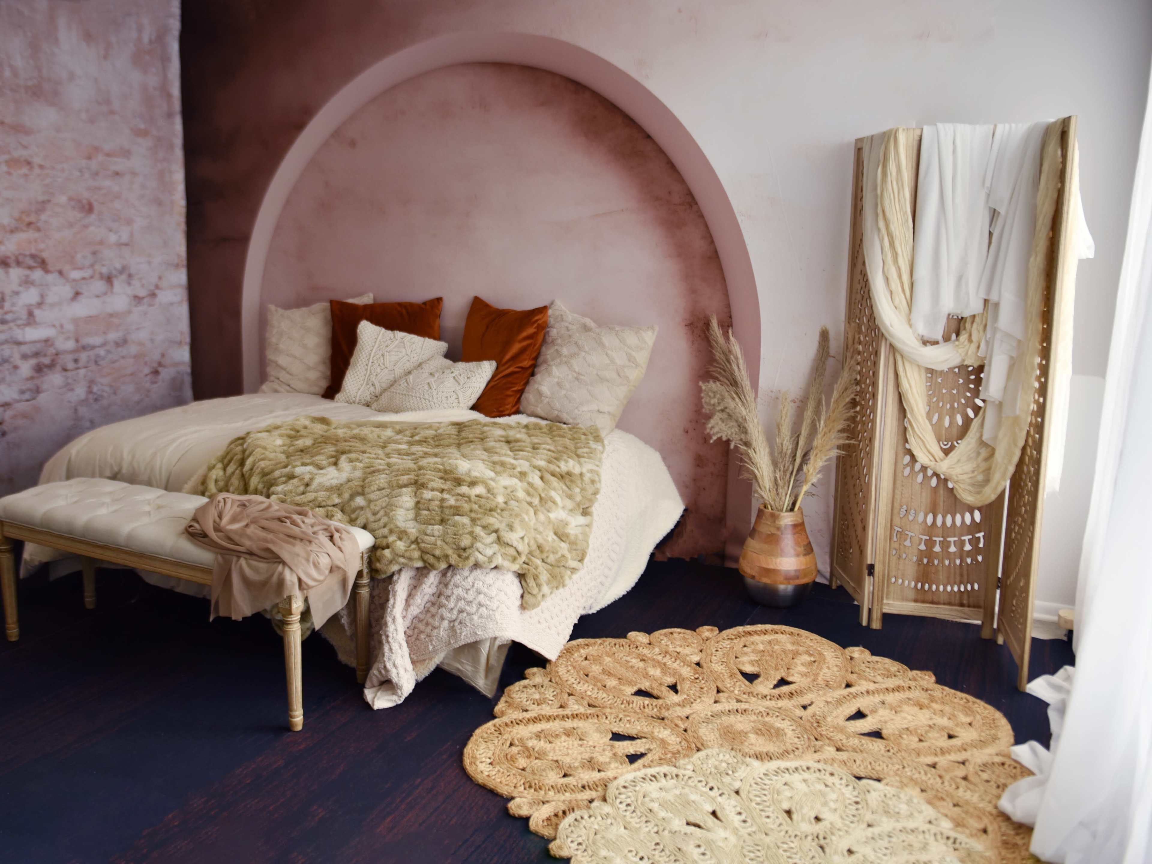 The image depicts a cozy bedroom featuring a textured wall backdrop, a bed adorned with various pillows and throws, a decorative rug, and a floor vase with dried plants.