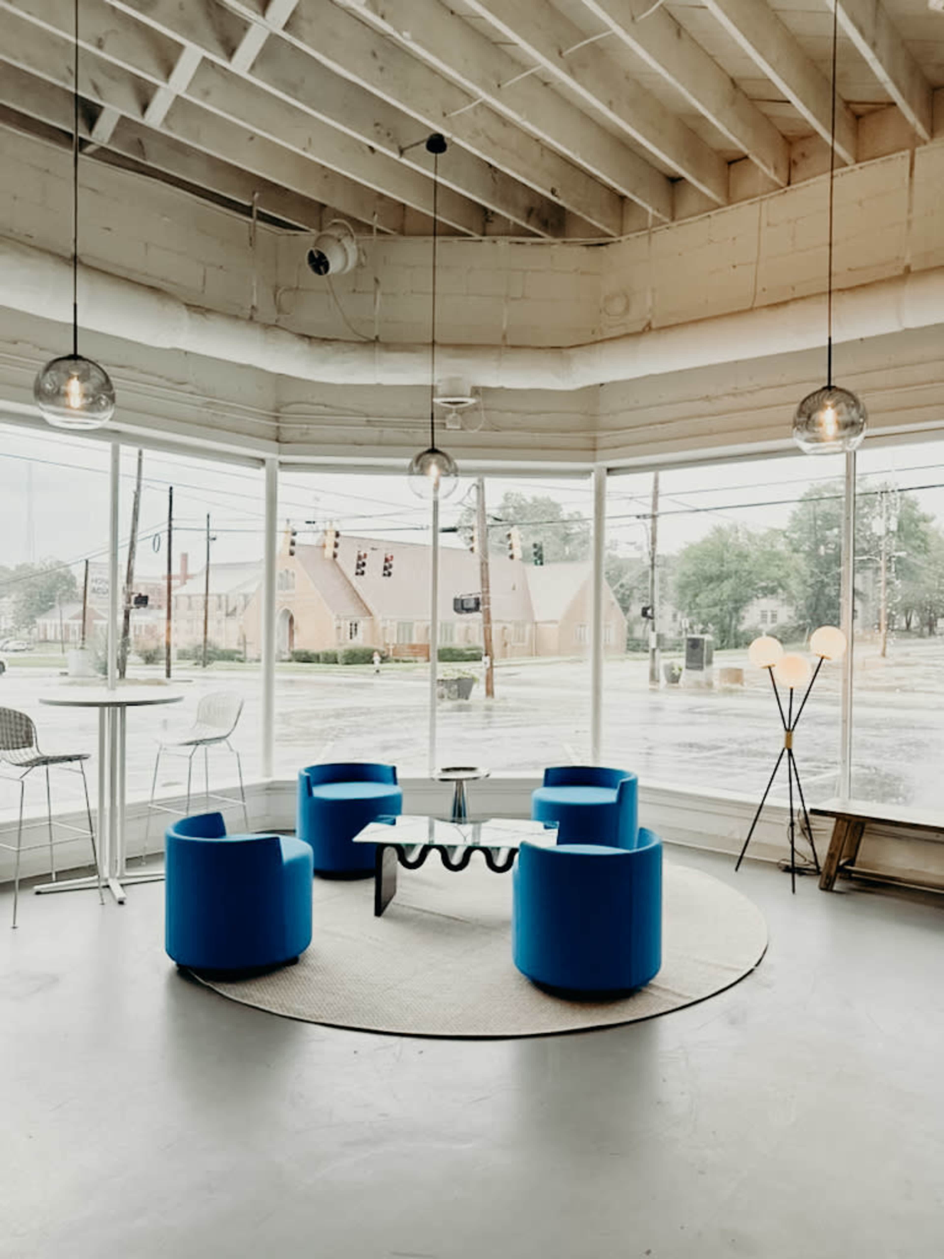 The image shows a modern interior with four blue chairs arranged in a circular formation around a coffee table, situated by large windows in a well-lit space.
