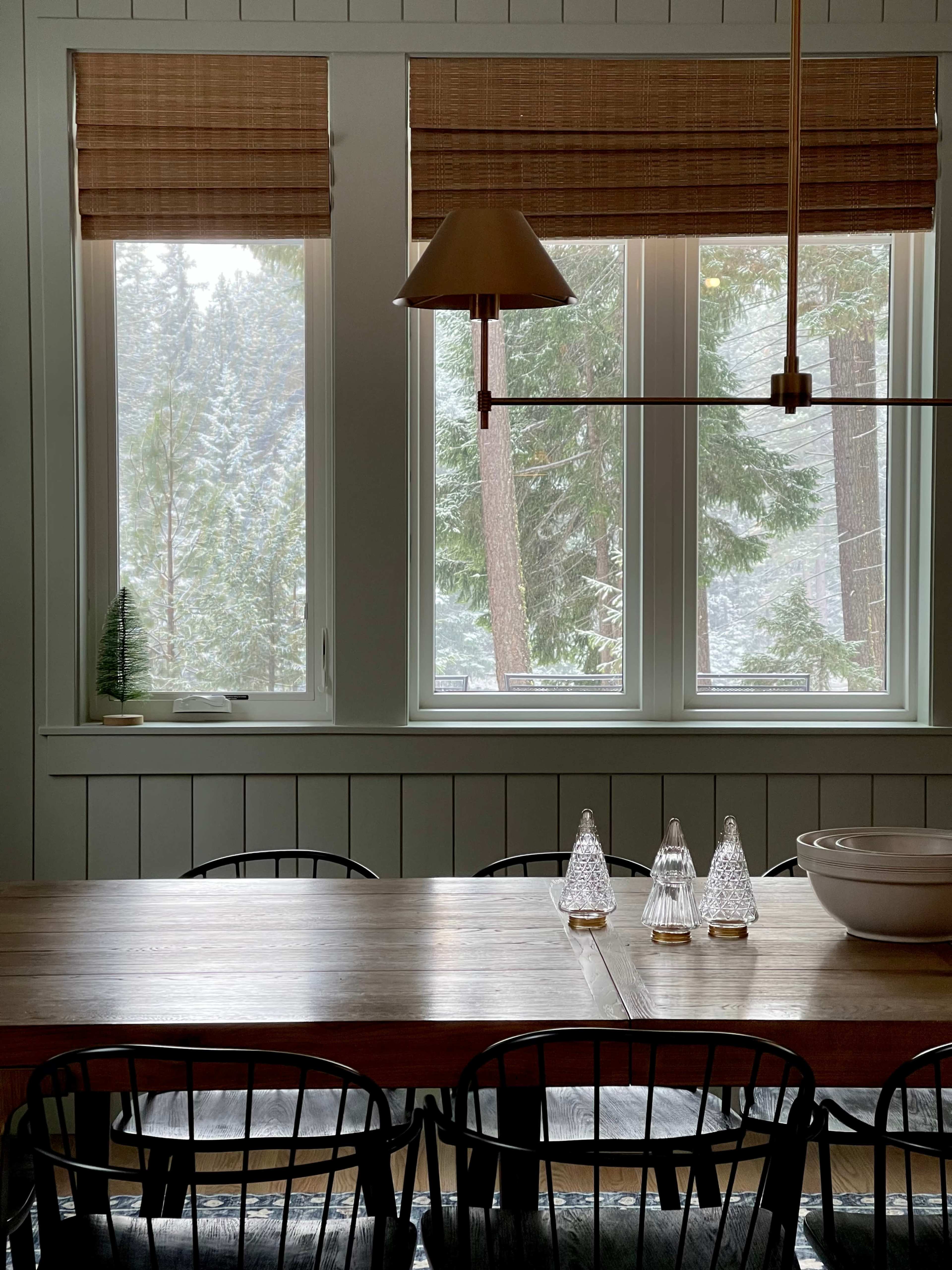 A wooden dining table with black chairs is set against a backdrop of large windows showing a snowy landscape and forest outside.