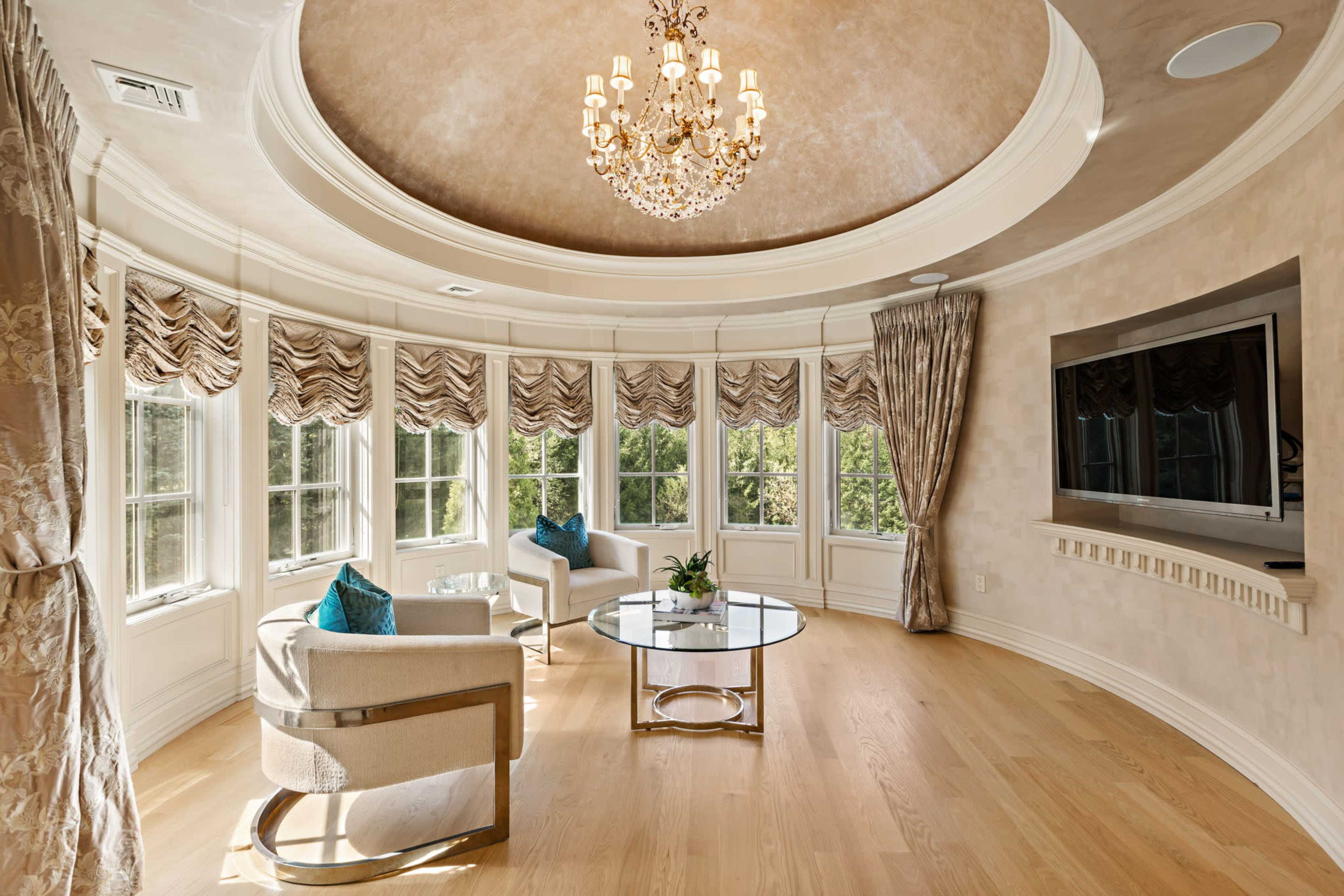 The image shows a circular room with large windows featuring patterned shades, a glass coffee table, two upholstered chairs, and a chandelier overhead.