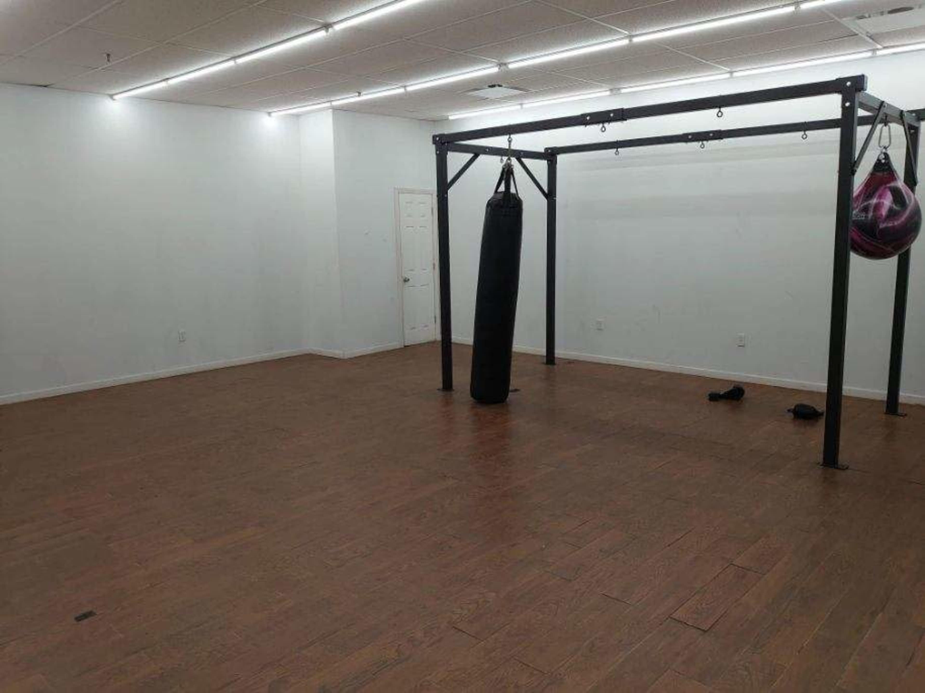 The image shows an empty exercise room with a wooden floor, featuring a heavy punching bag hung from a black steel frame.