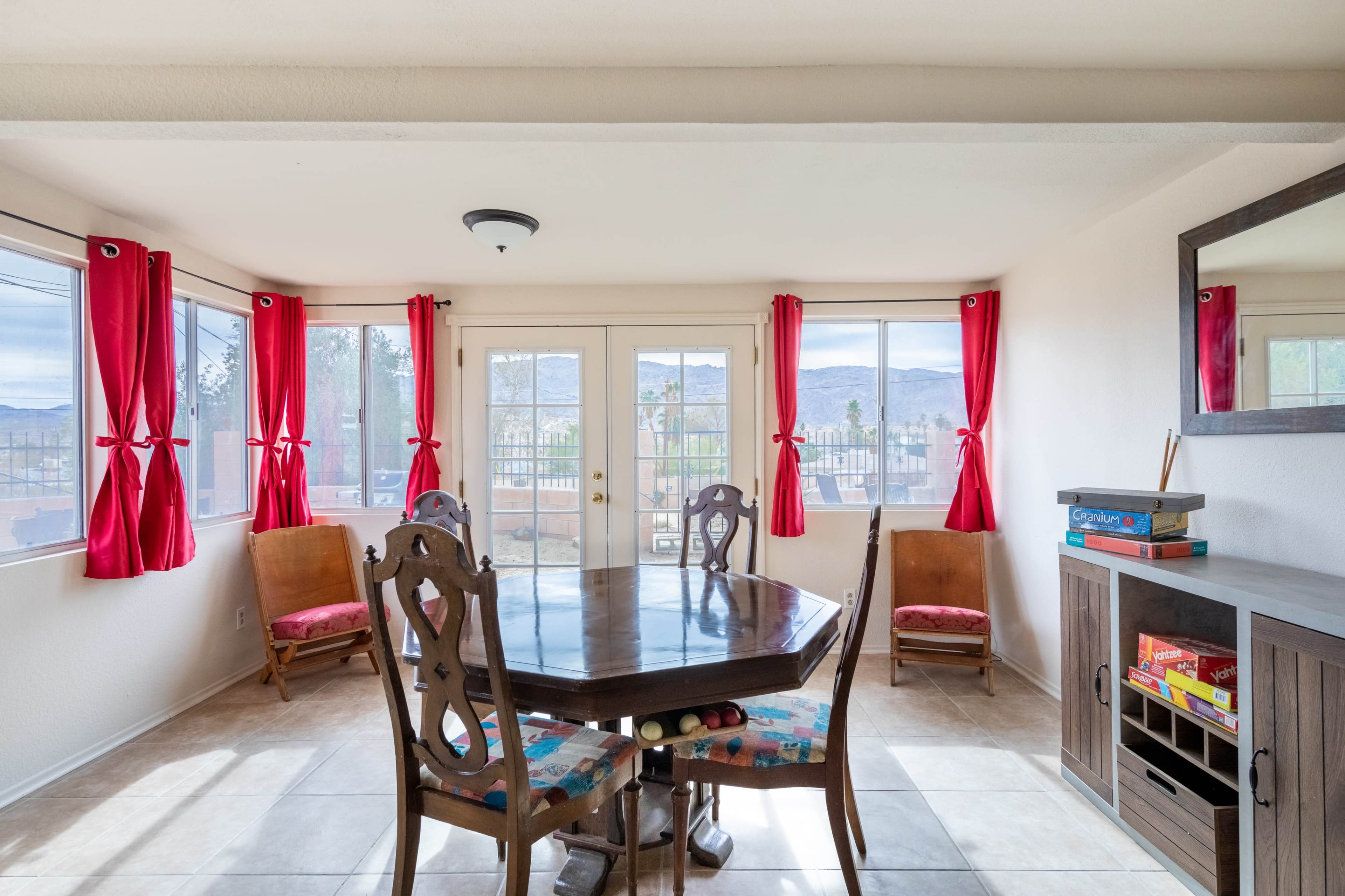 A dining area features a round table surrounded by chairs, with large windows dressed in red curtains and a view of the outdoors.