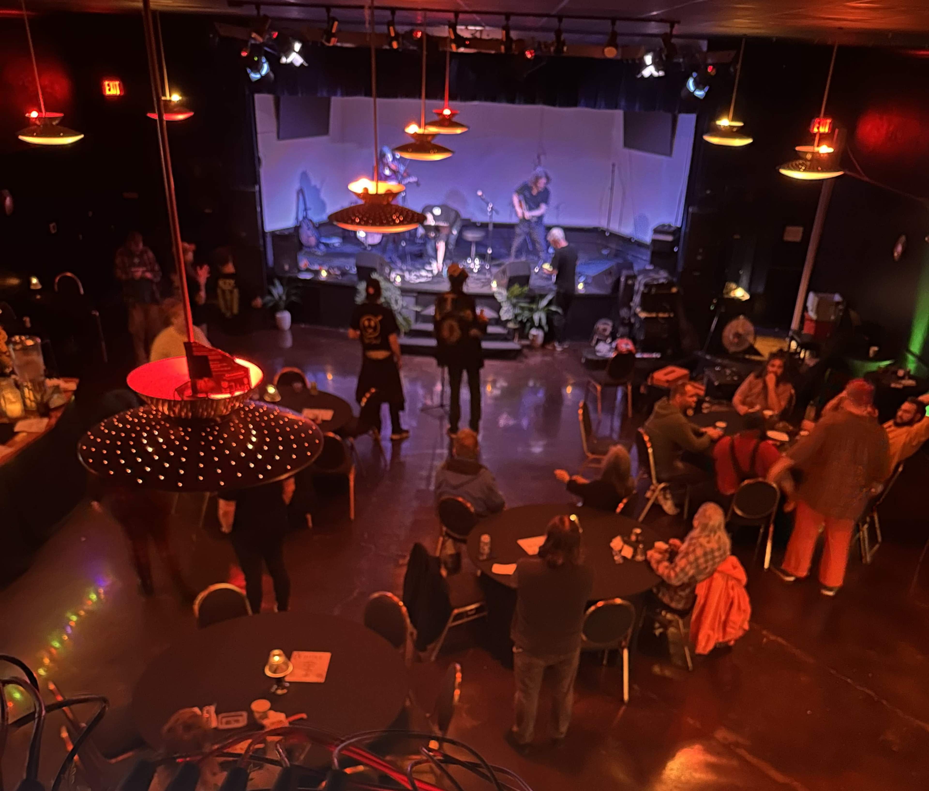 A live music venue features a stage set up with instruments, while attendees gather at tables and the bar area.