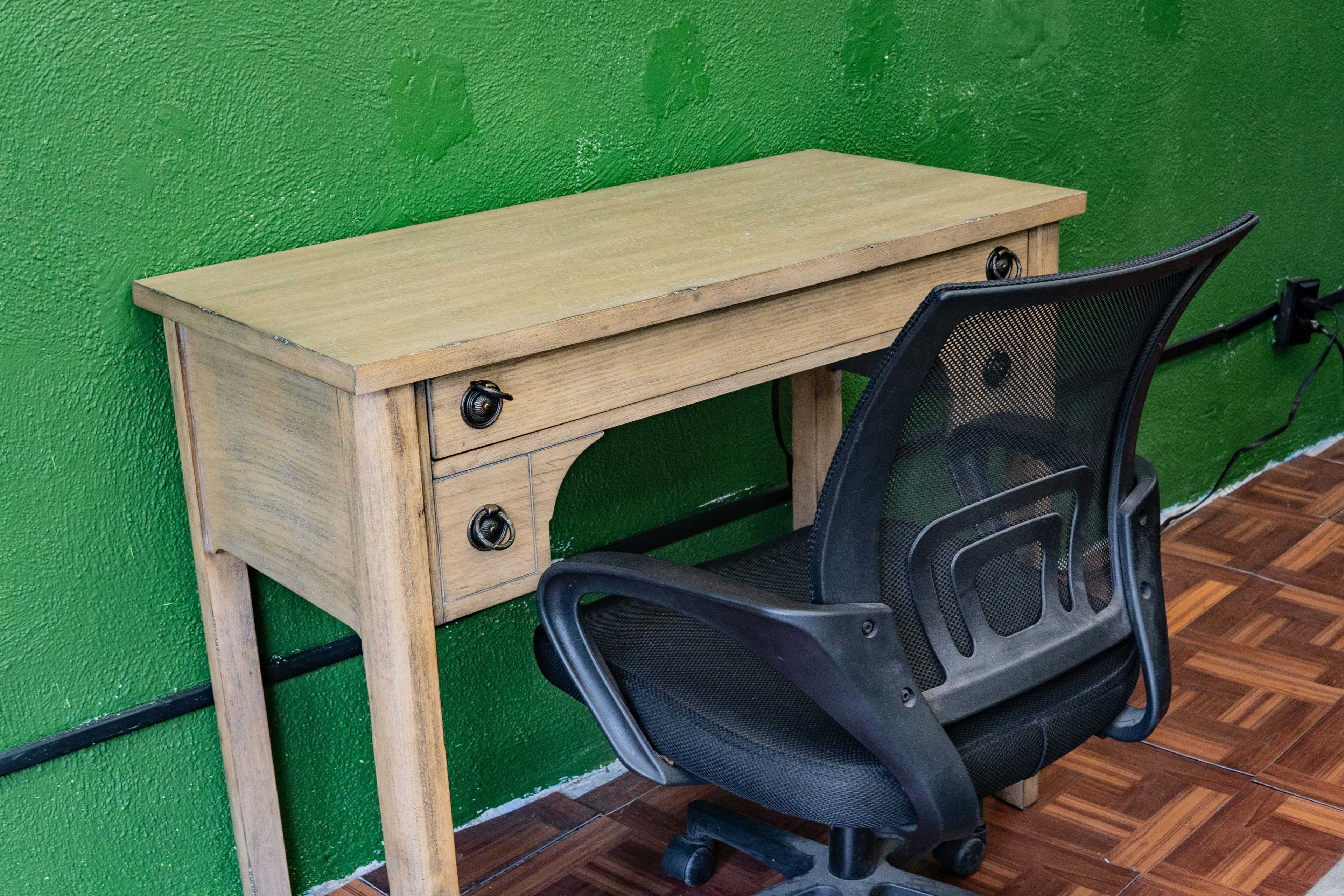 A wooden desk with two drawers is positioned against a green wall, accompanied by a black office chair.