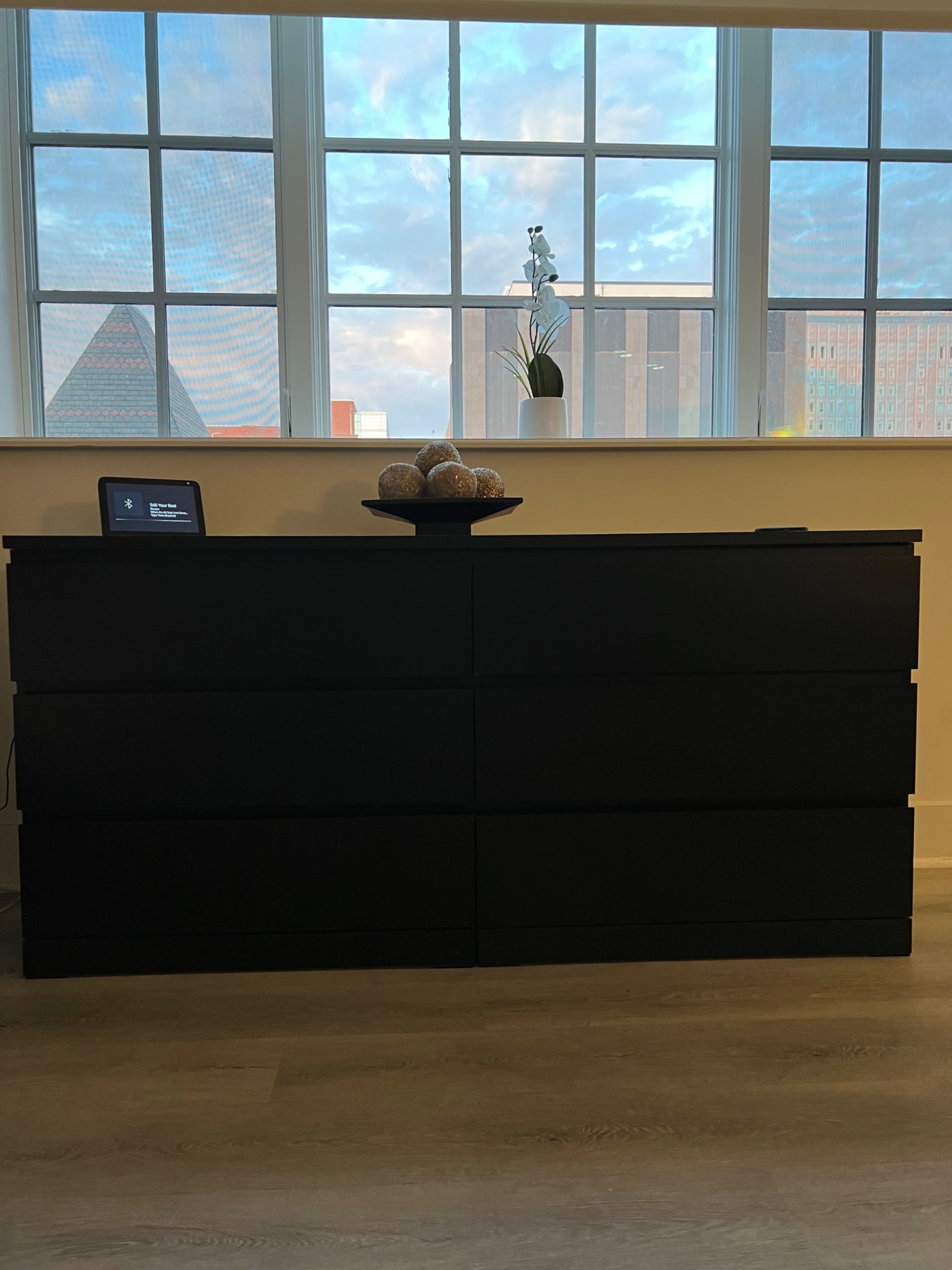 A black dresser with six drawers stands in front of large windows, which frame a skyline view and allow natural light to enter the room.