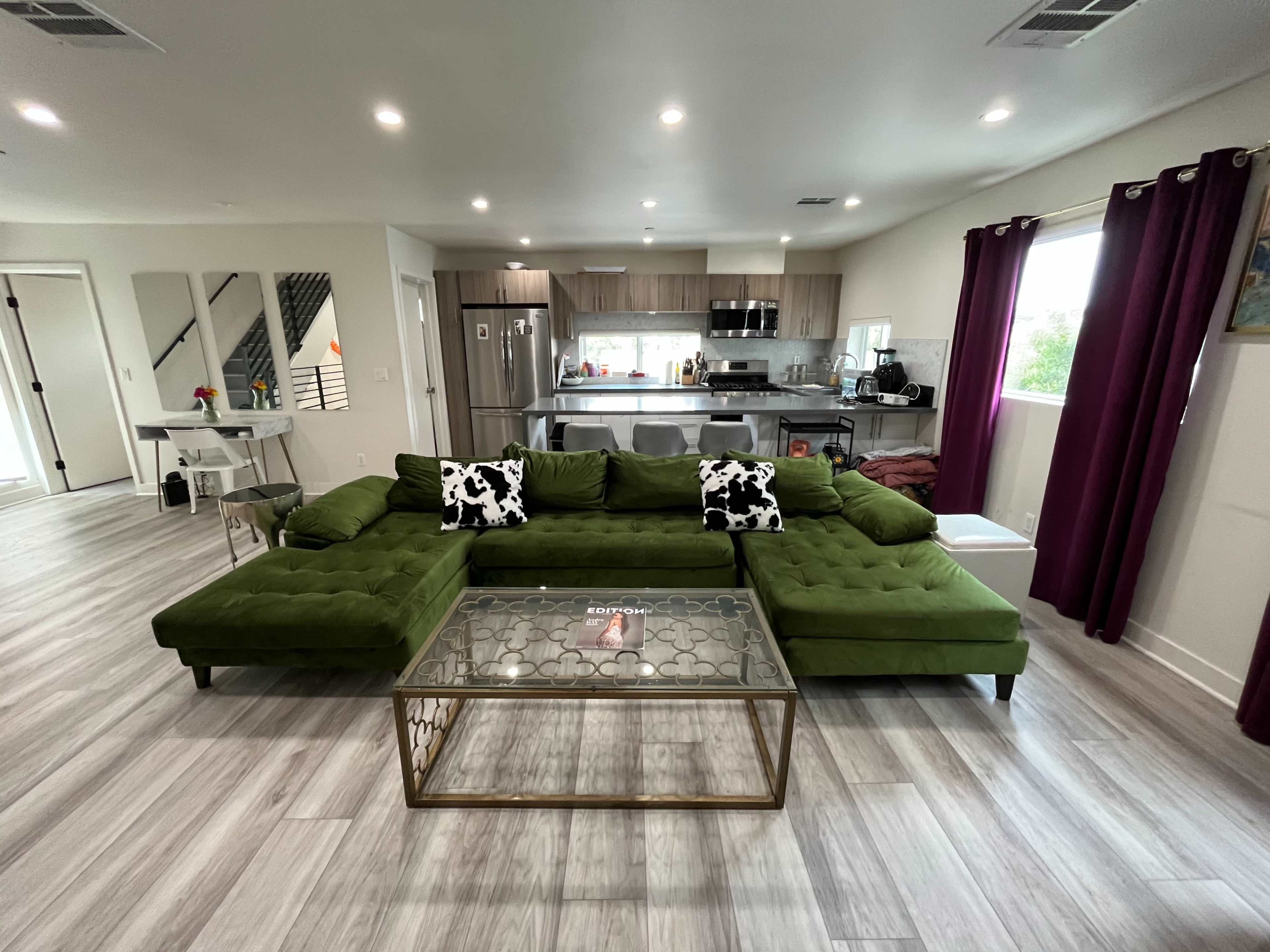 The image shows a modern living room with a green sectional sofa, a glass coffee table, and an open kitchen in the background.