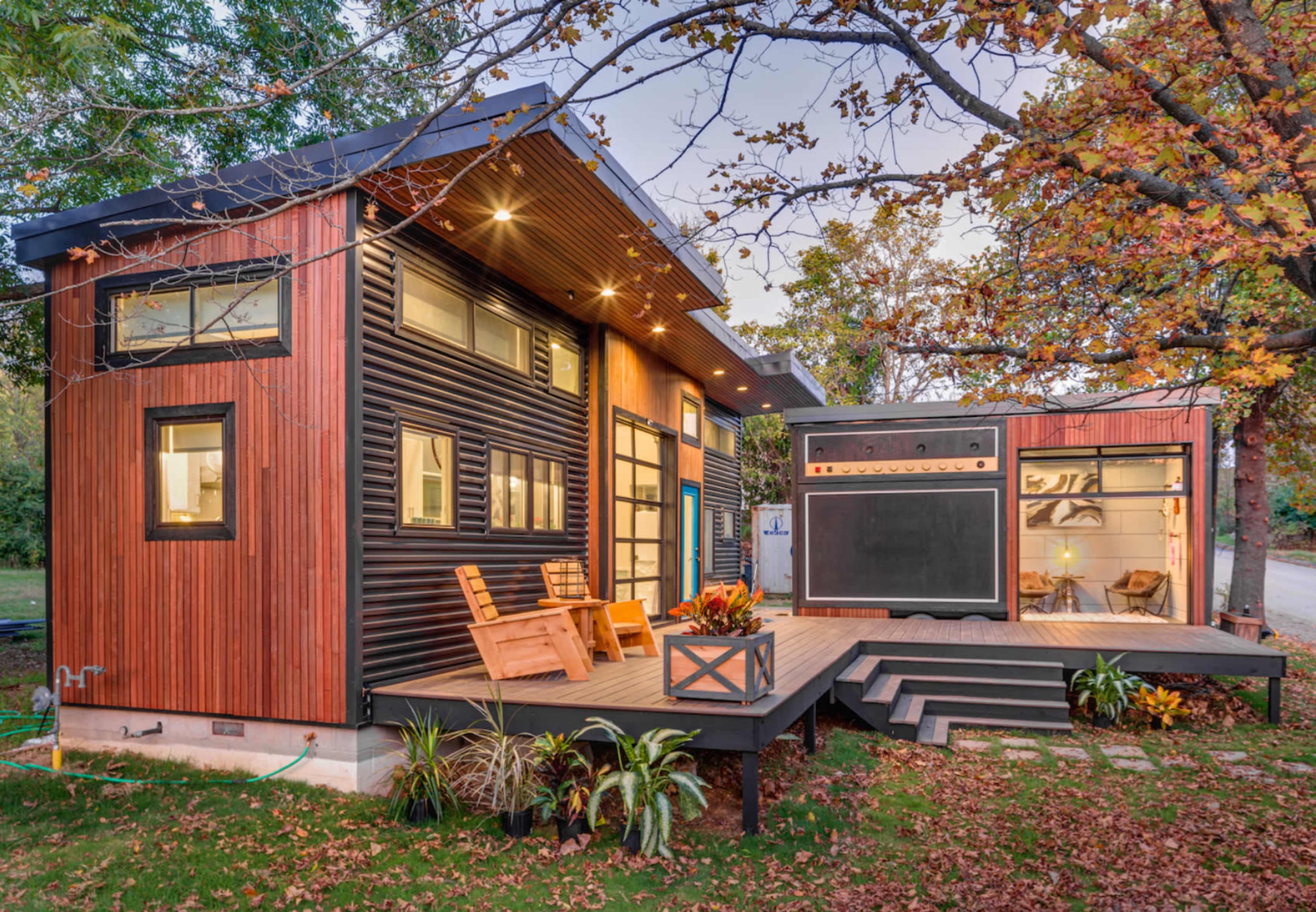 A modern house with a wooden deck and two outdoor chairs, surrounded by trees in autumn foliage.