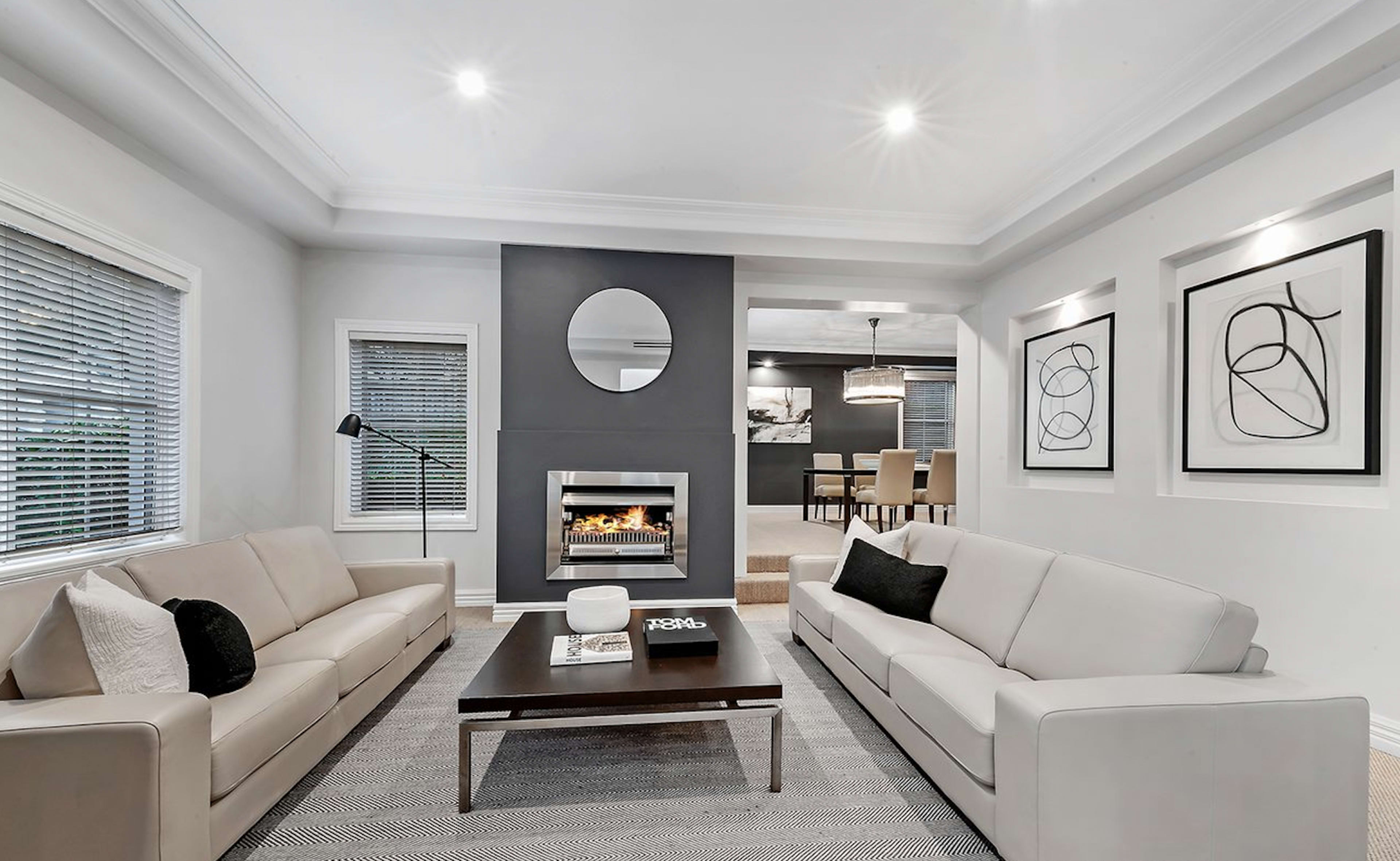 The living room features two light-colored sofas facing a central coffee table, with a fireplace and a circular mirror on the wall in the background.