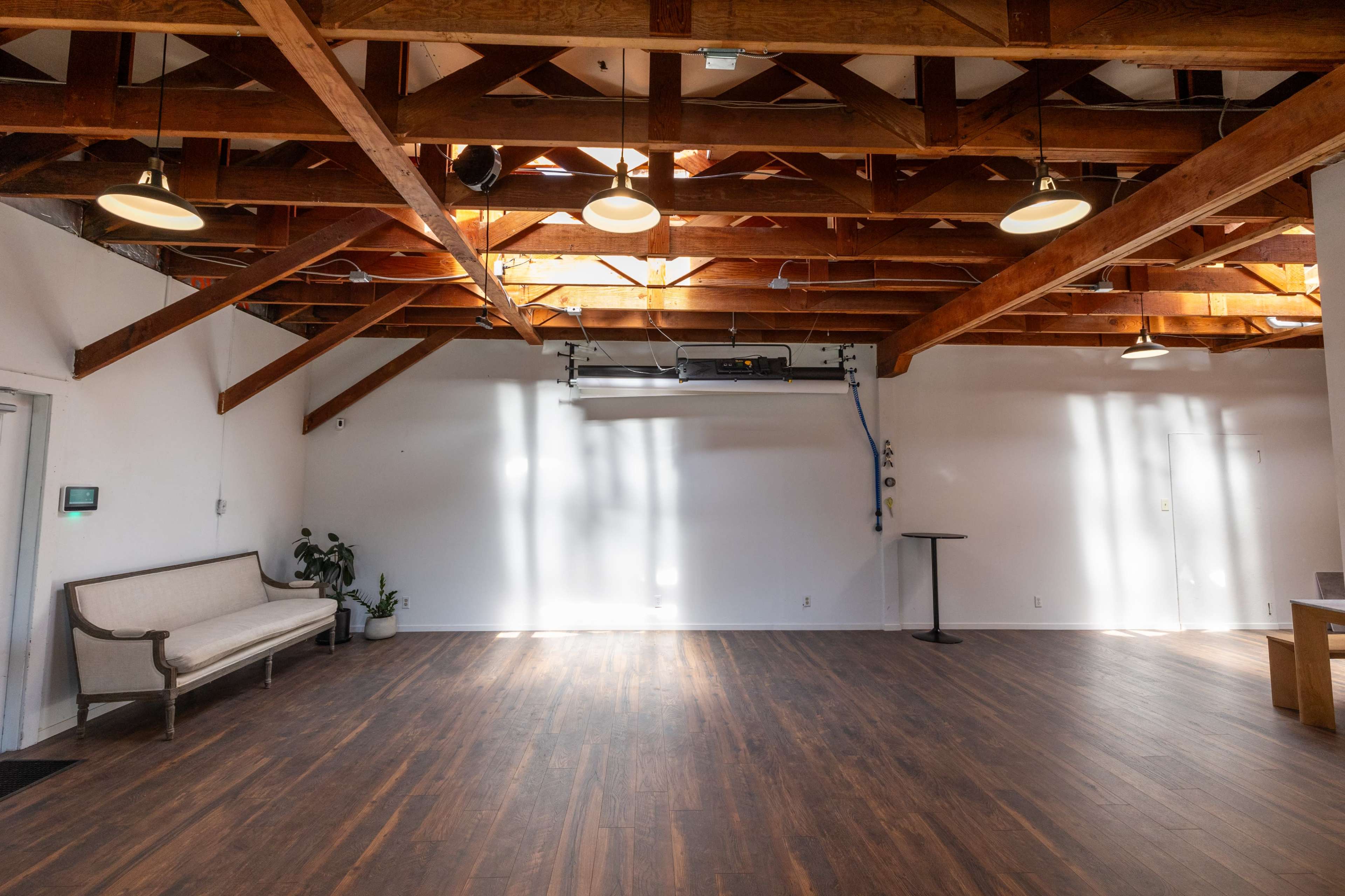 A spacious, well-lit room with wooden beams, hardwood flooring, and minimal furniture including a bench and a small table.