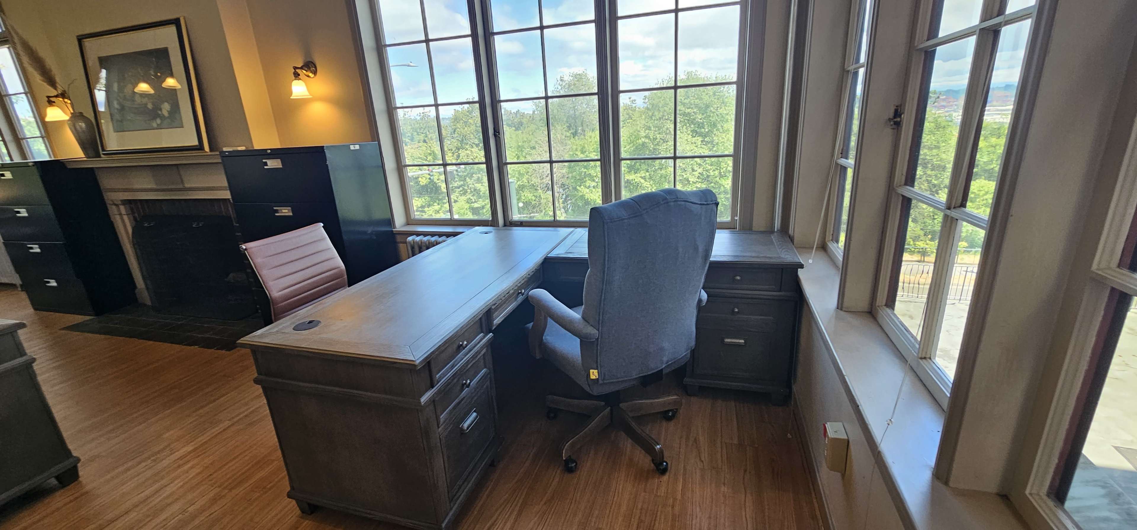 The image shows a spacious office interior with large windows, a wooden desk, a rolling chair, and file cabinets against the walls.