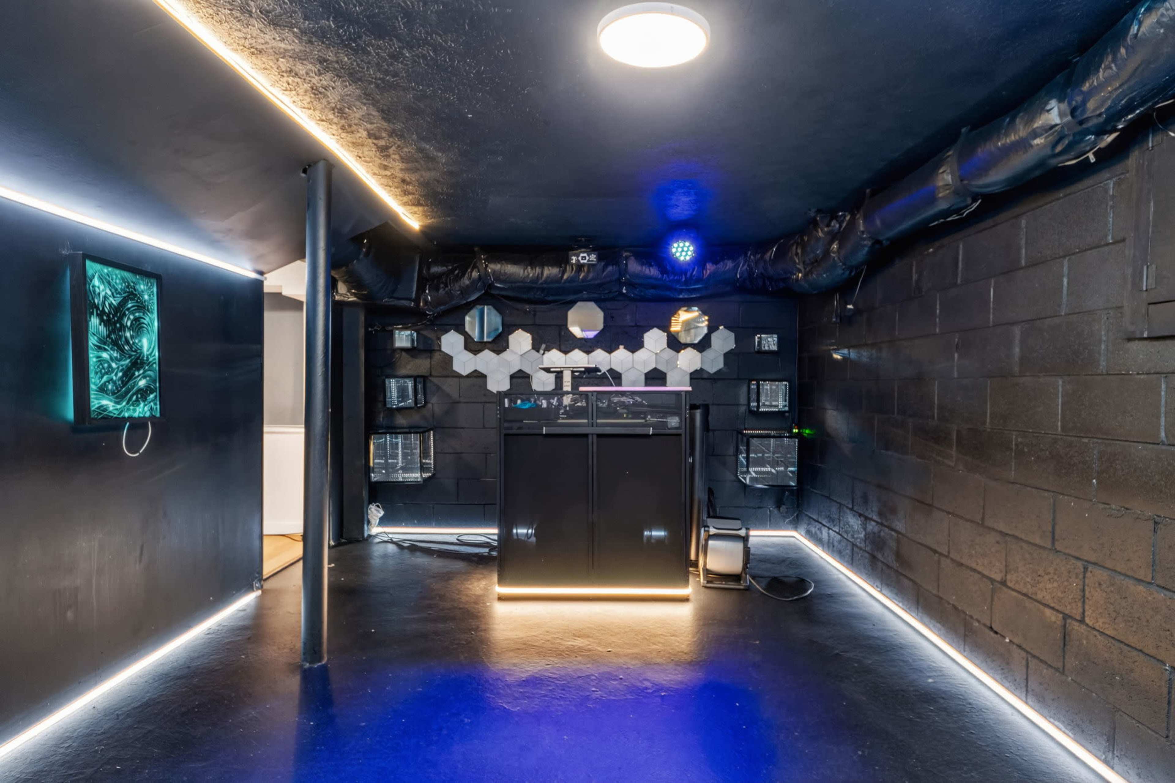 The image shows a dimly lit, modern room with a black aesthetic featuring LED lighting, a DJ setup against the back wall, and various monitors mounted on the adjacent walls.