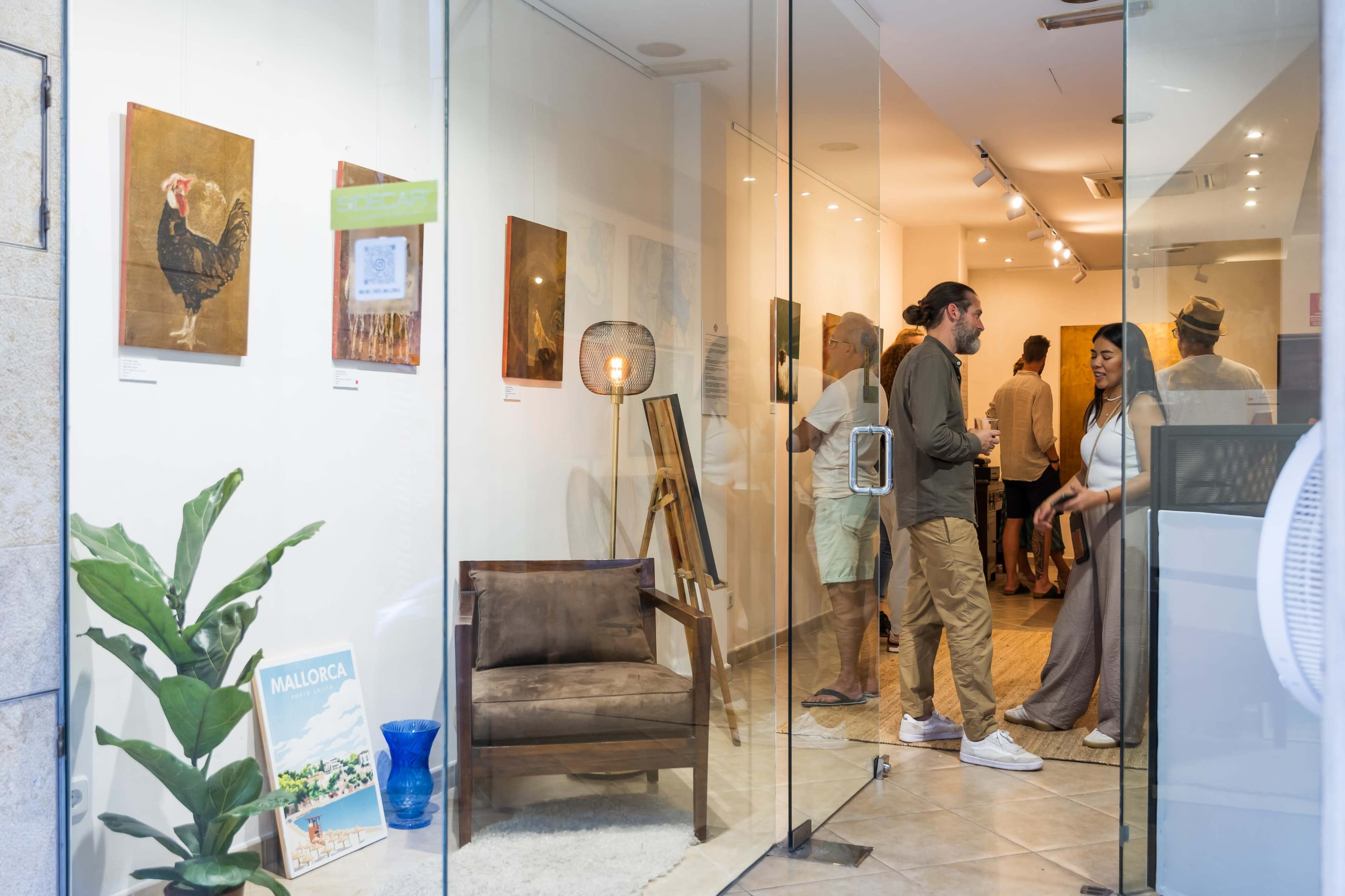 The image shows a gallery setting with several people engaging in conversation, surrounded by artworks displayed on the walls.