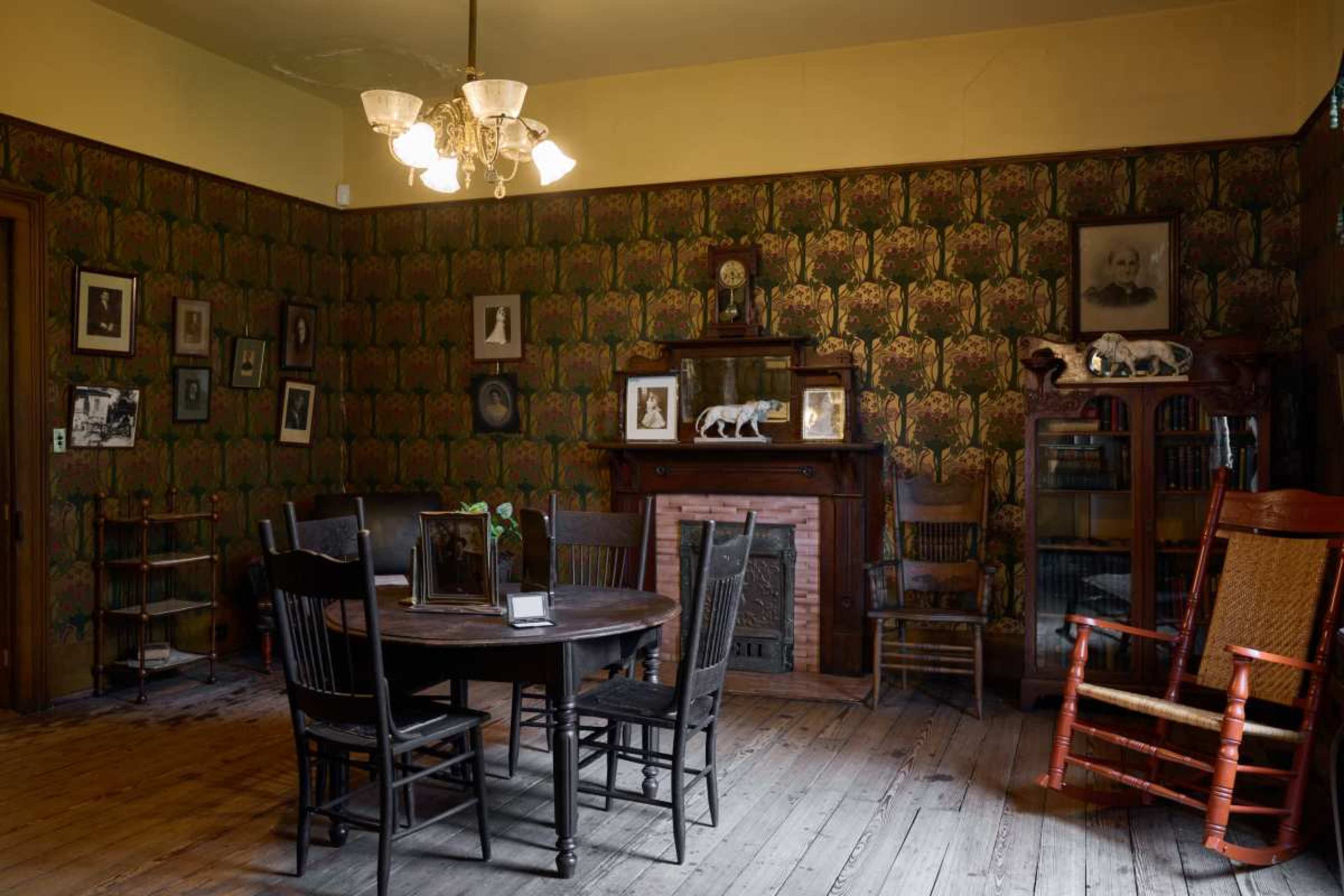 The image shows a vintage room featuring a round table surrounded by wooden chairs, a rocking chair in the corner, and walls adorned with patterned wallpaper and framed photographs.