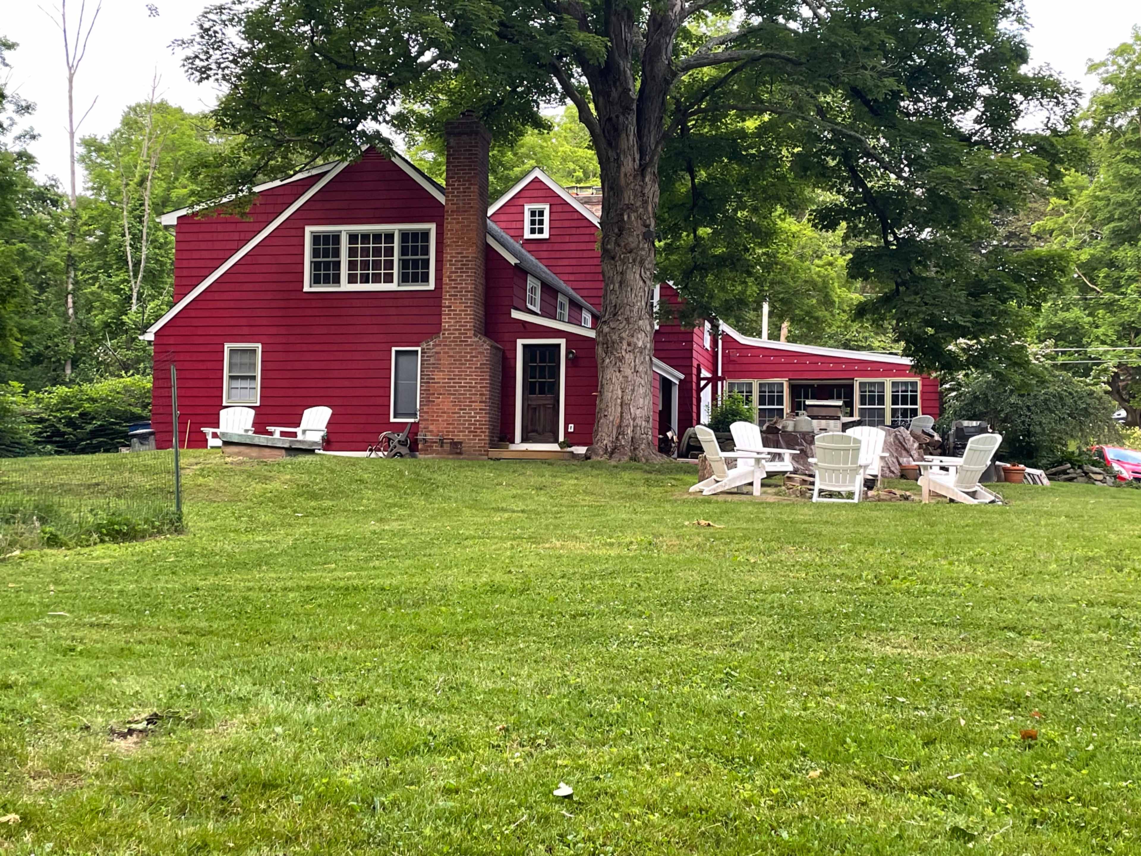 A red two-story house with a chimney and a porch, surrounded by a grassy yard and several white lawn chairs.