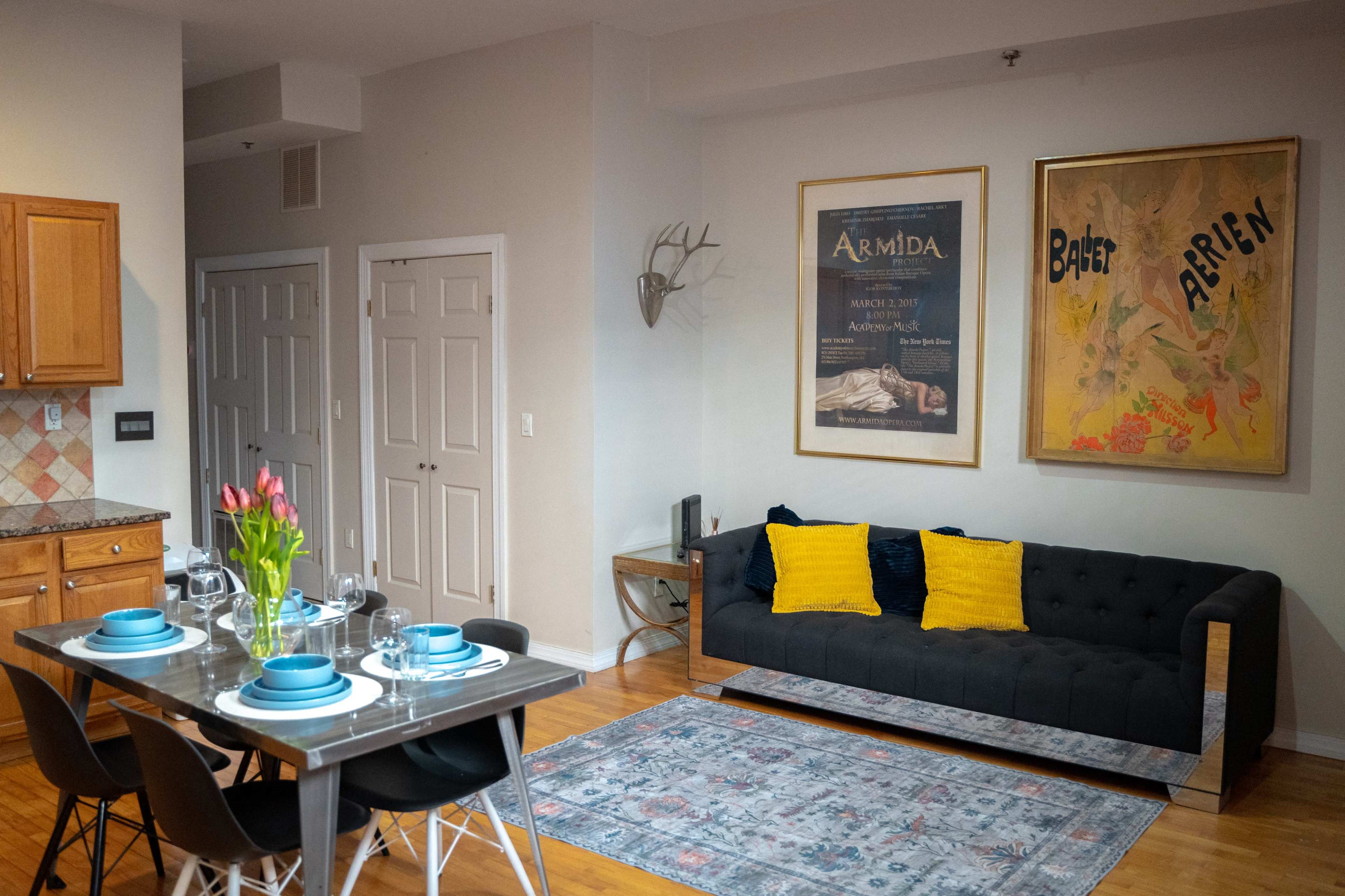 The image shows a modern living space featuring a dark sofa with yellow cushions, a dining table set for dinner, and framed art on the walls.