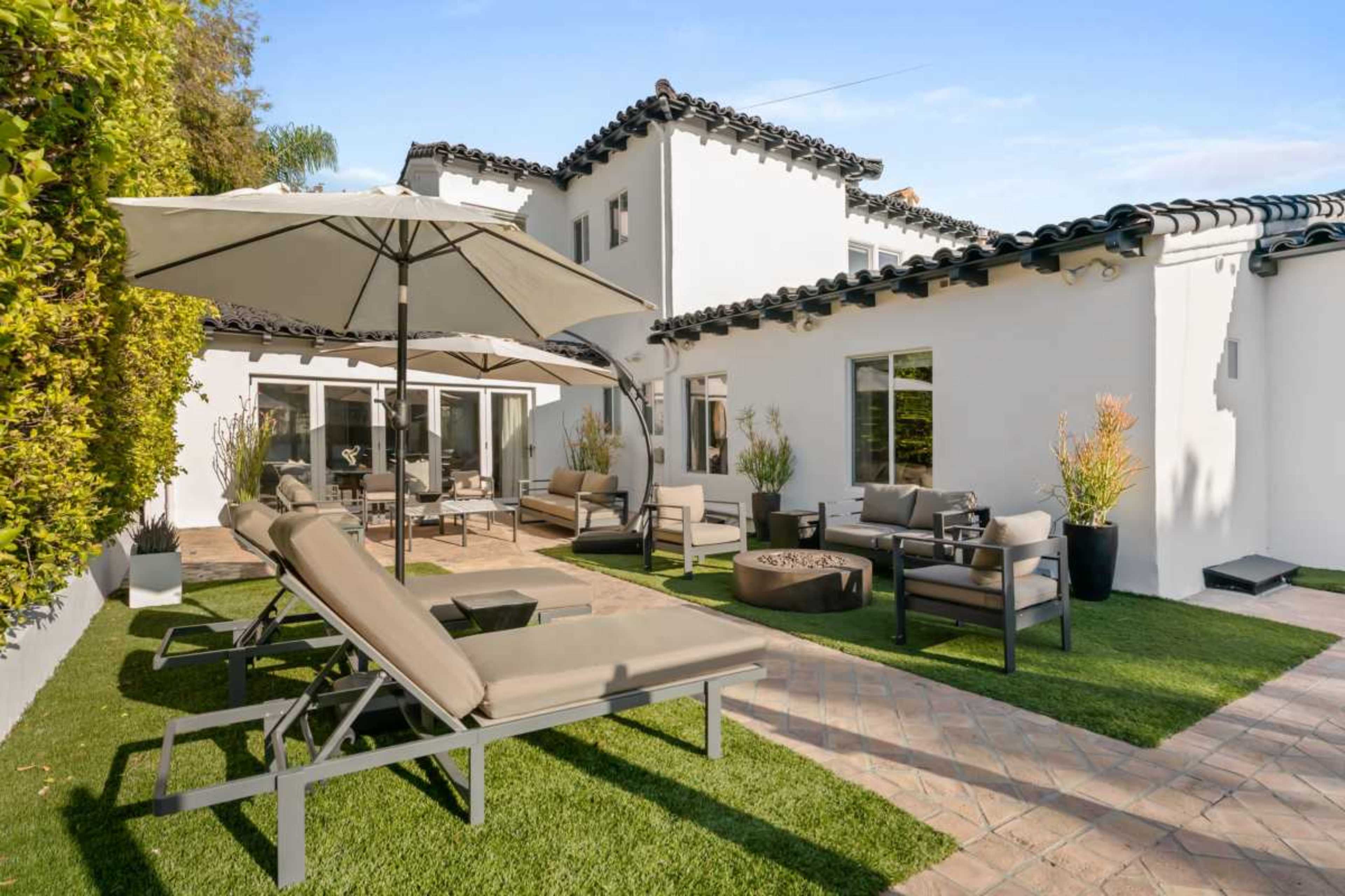 The image shows a modern outdoor patio area featuring lounge chairs, an umbrella, and seating arrangements next to a well-maintained lawn and a white house with a tiled roof.