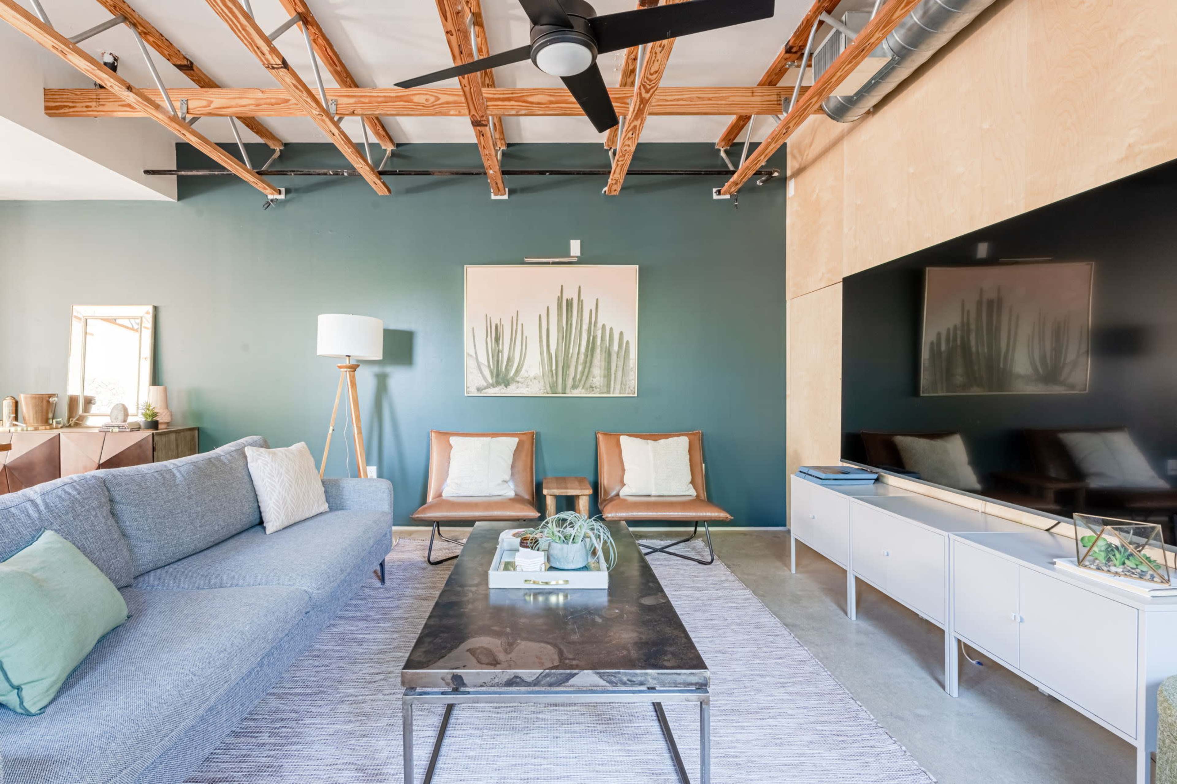 A modern living room features a gray sofa, two leather chairs, a coffee table, and a wall-mounted TV, with decorative elements like a plant artwork and a lamp.