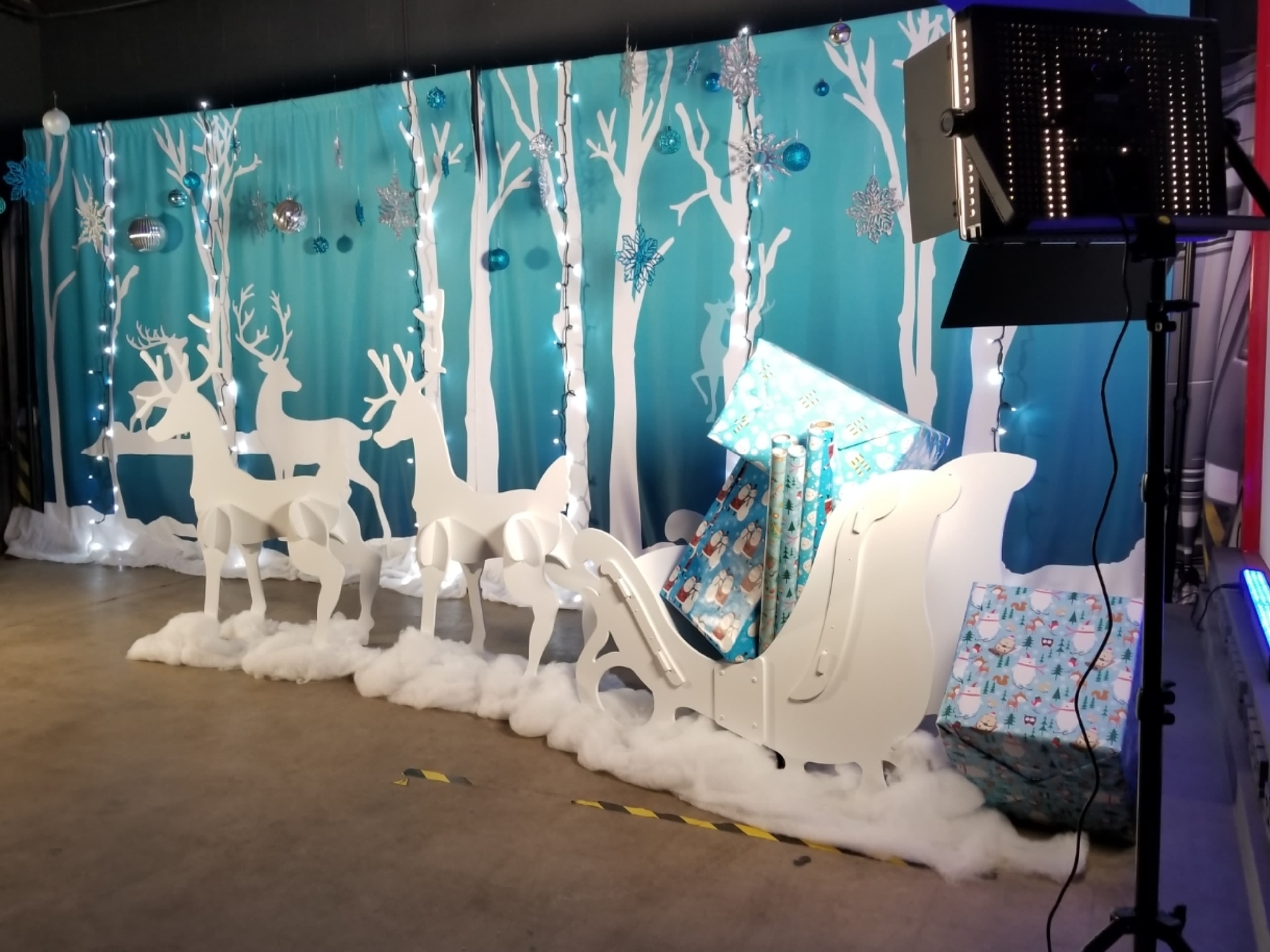 A festive display features a white sleigh with wrapped gifts, surrounded by white reindeer silhouettes and decorated snowy trees against a blue backdrop.