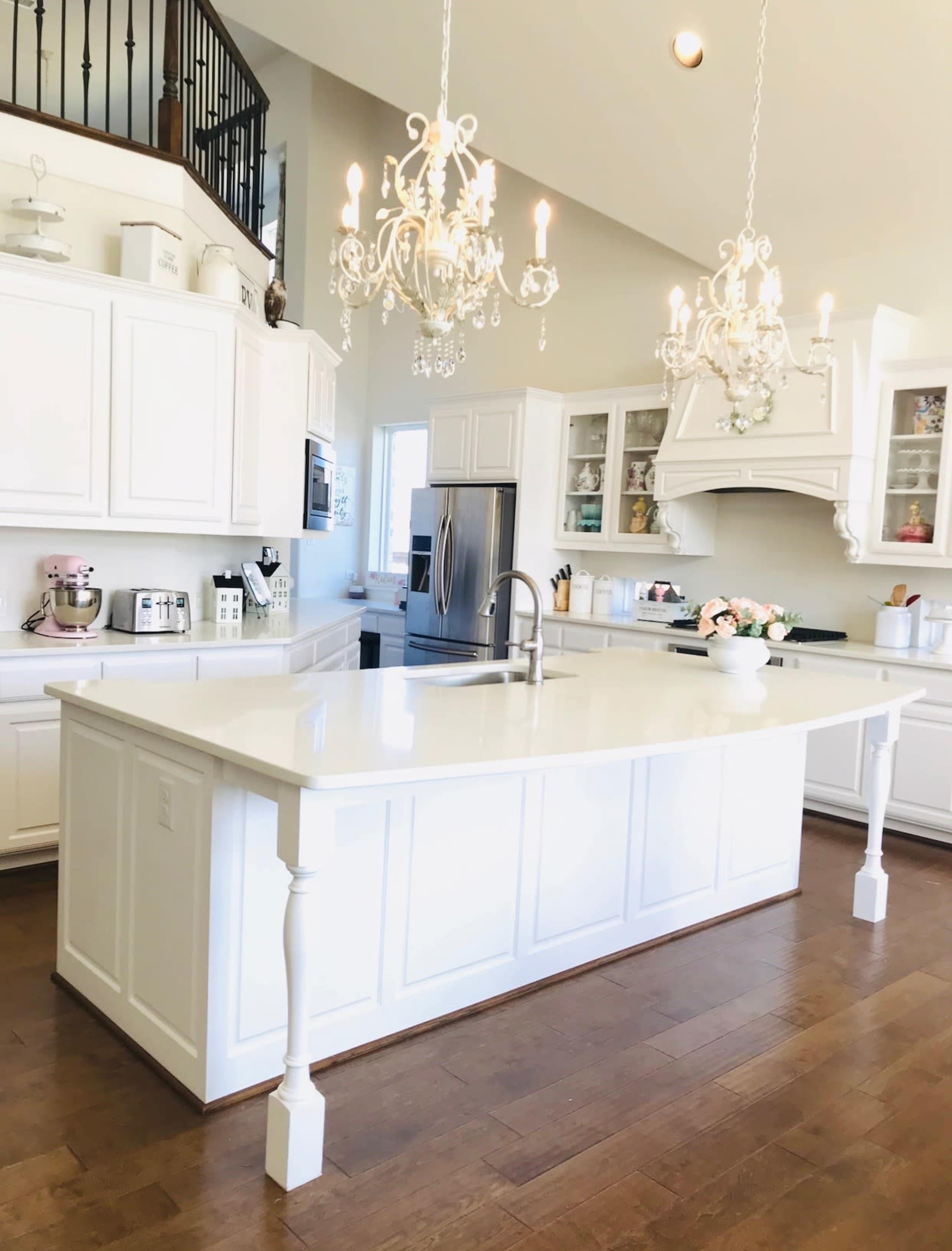 A modern kitchen with white cabinetry, an island with a white countertop, and two elegant chandeliers hanging from the ceiling.