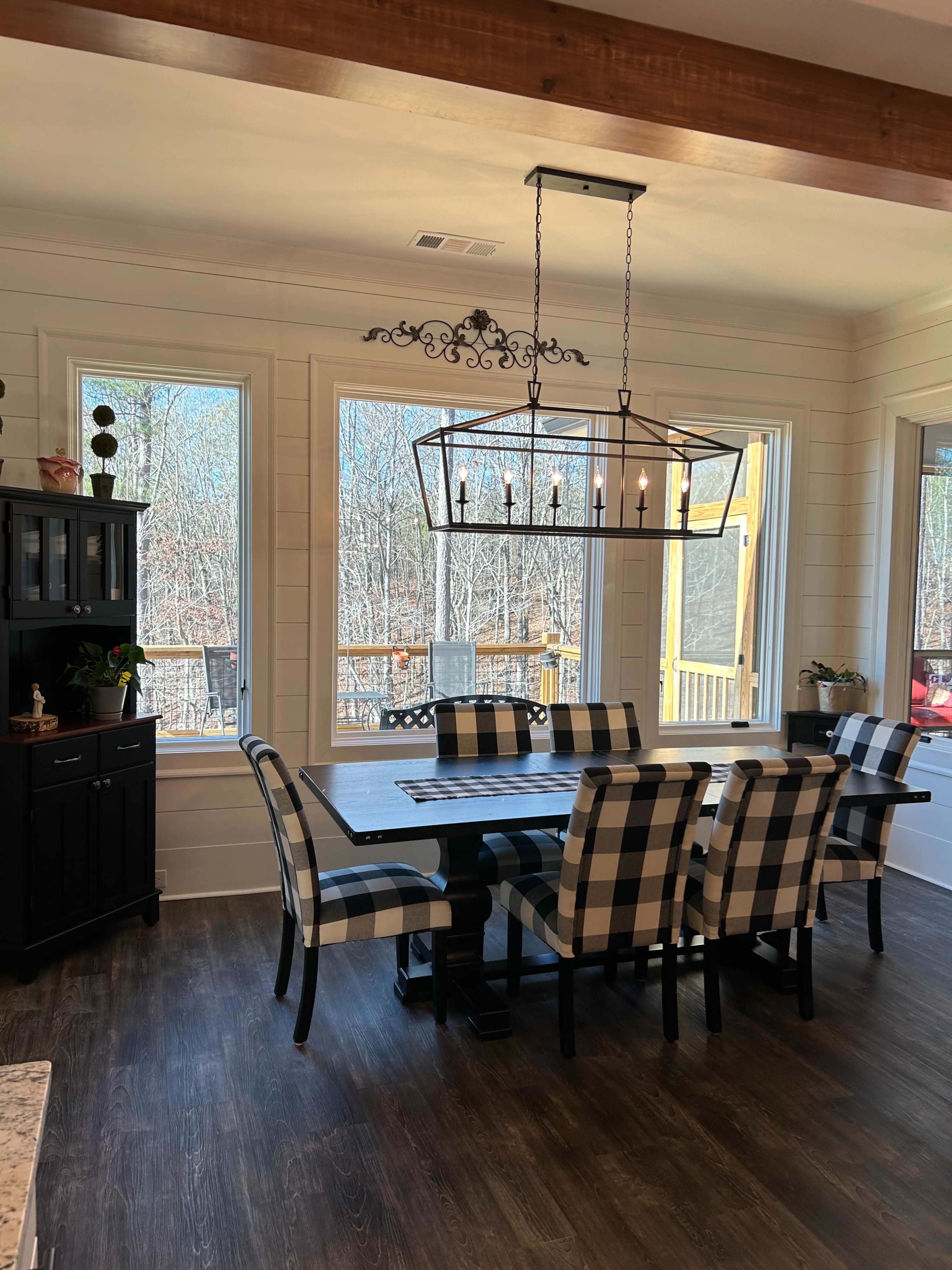 A dining area features a large checkered table with matching chairs, surrounded by windows that provide a view of the wooded exterior.