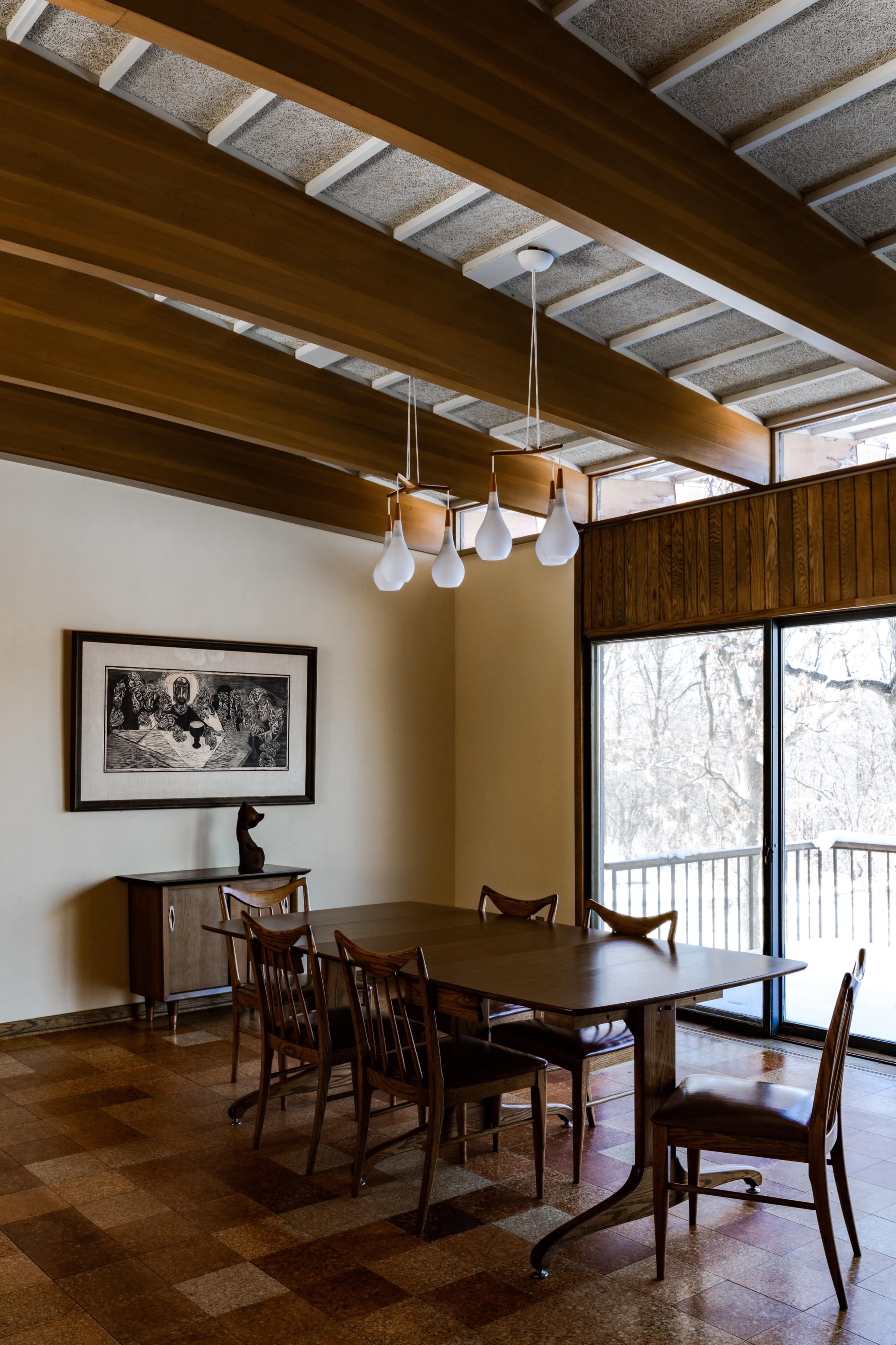 The image shows a modern dining room with wooden beams, a large table surrounded by chairs, and a framed artwork on the wall near a window with views of a snowy landscape outside.