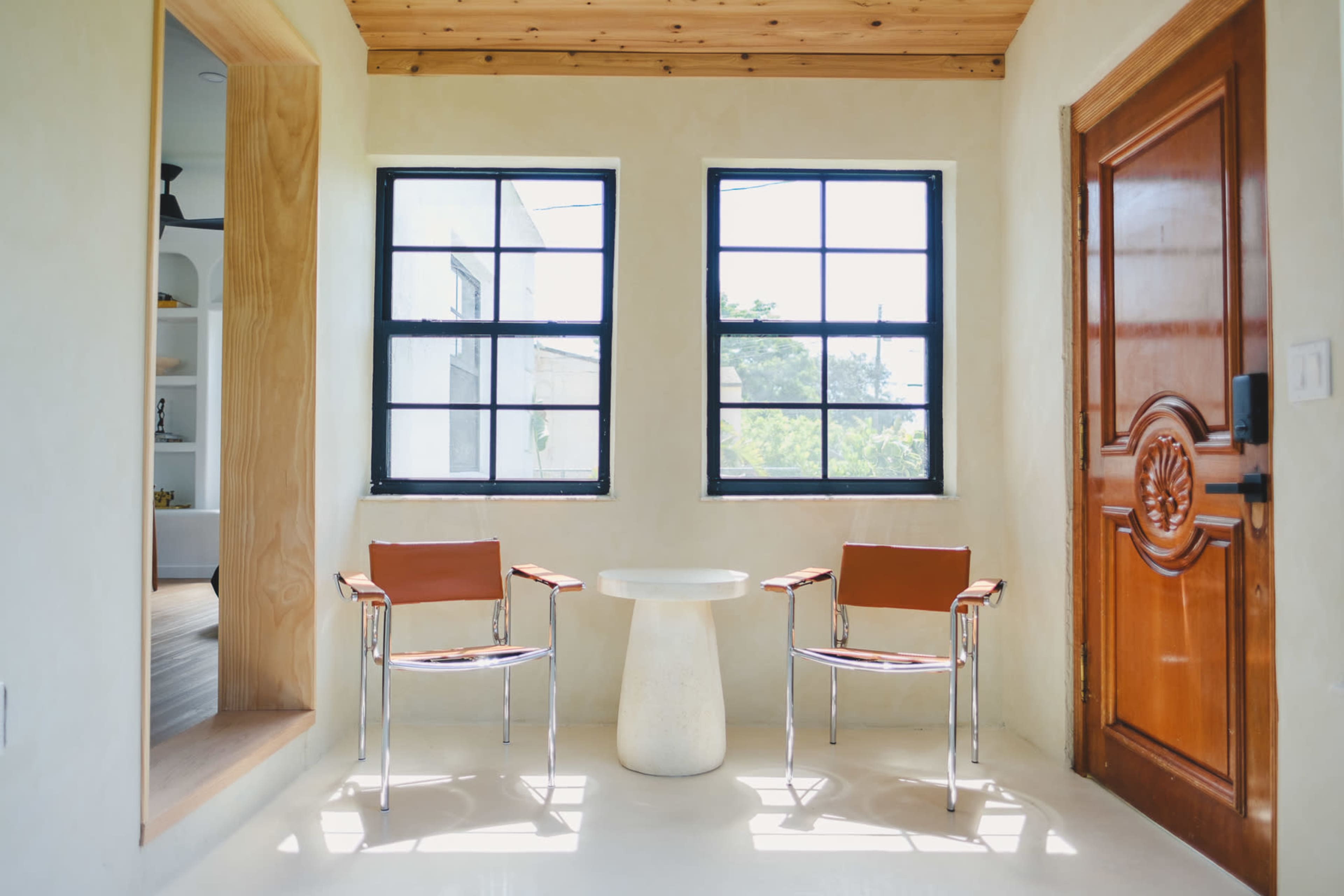 The image shows a bright, minimalistic room with two chairs and a round table, complemented by wooden accents and large windows.