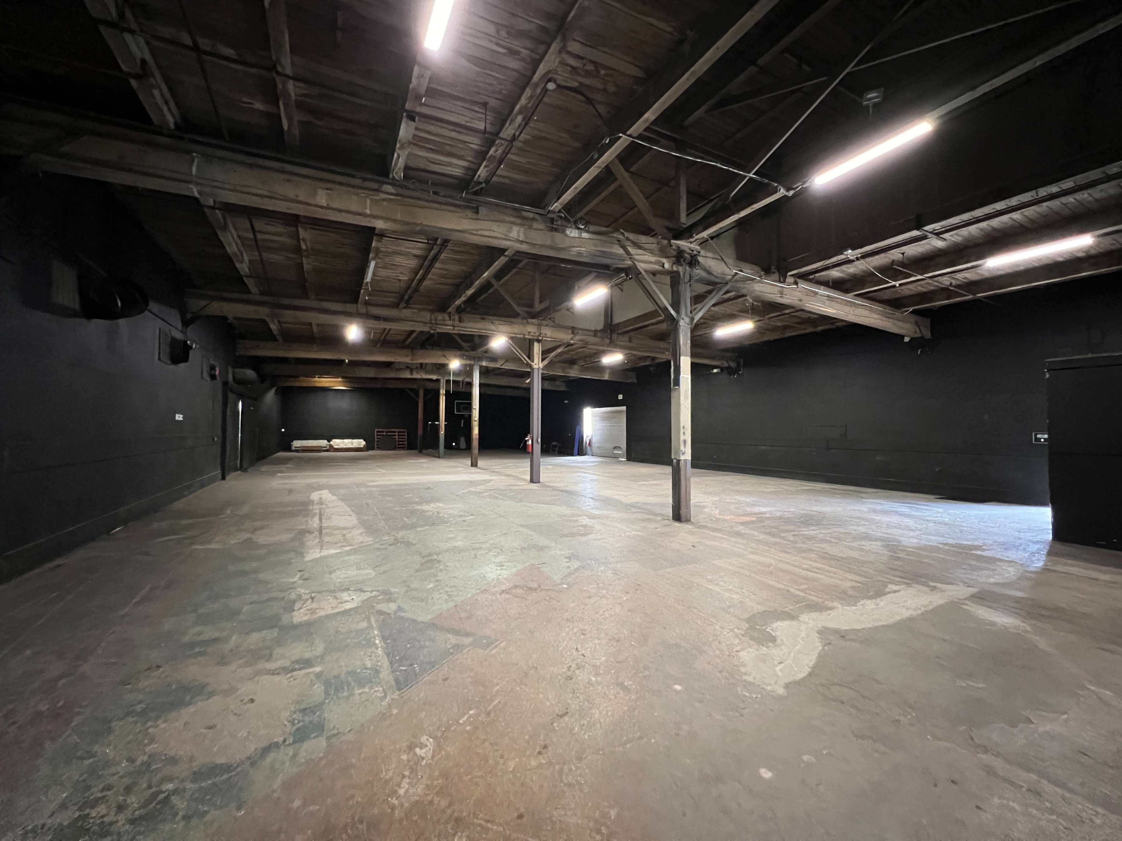 A large, empty indoor space with a wooden ceiling and concrete floor, illuminated by fluorescent lights.