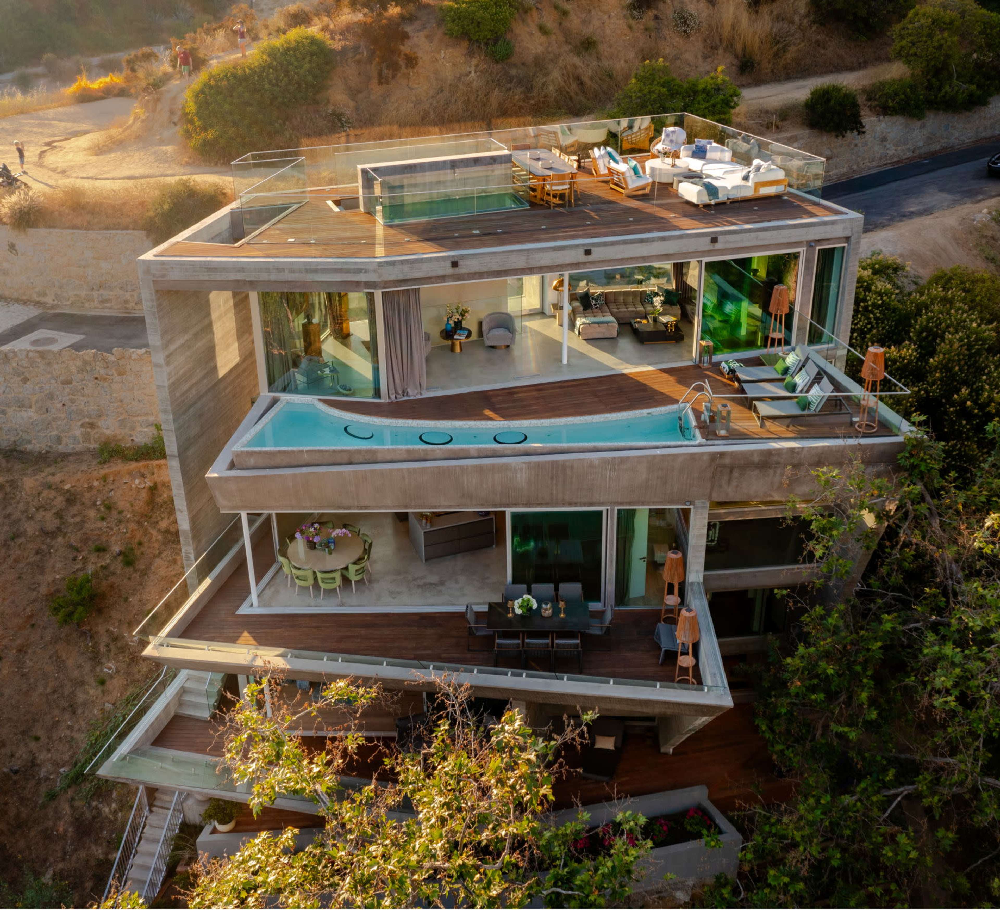 A modern multi-level house with a large glass facade, an infinity pool on the terrace, and outdoor seating areas surrounded by greenery.