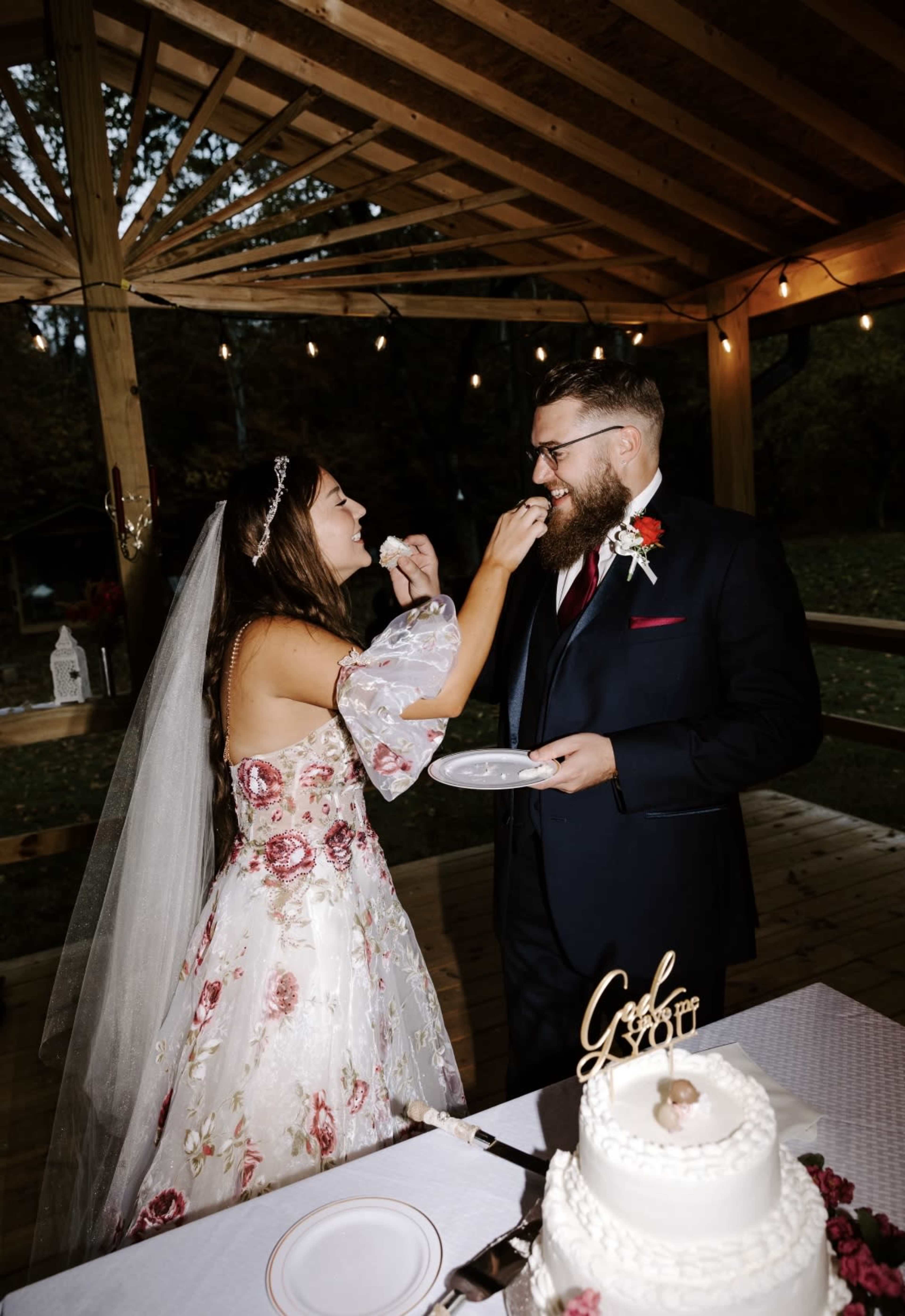 A bride in a floral dress feeds cake to a groom at an outdoor wedding reception under a wooden pavilion.